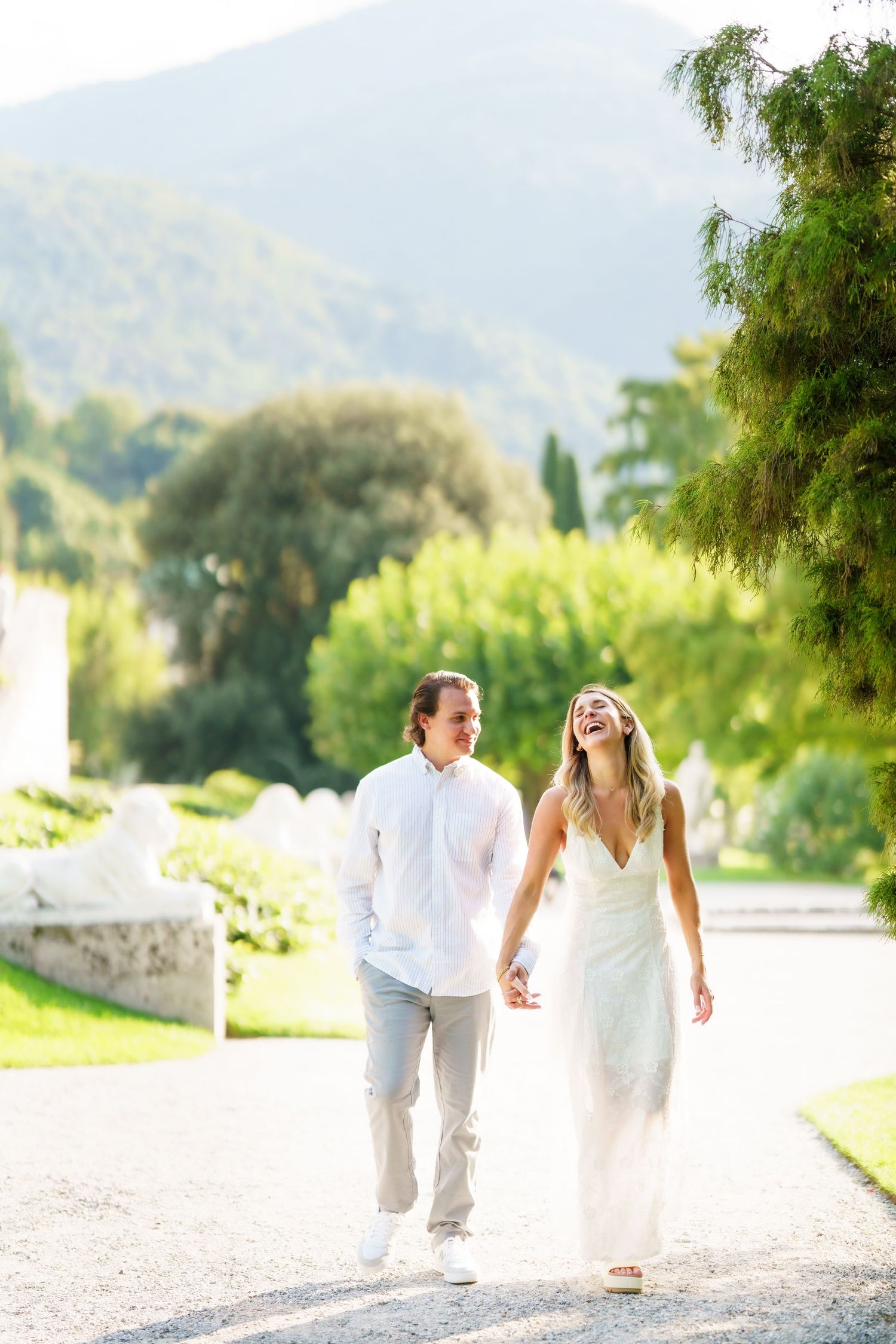 A couple walks and laughs together on a sunlit path surrounded by lush greenery and mountains in the background.