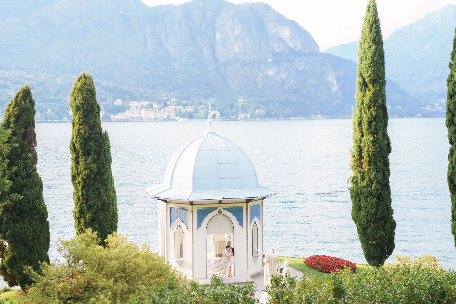Tall cypress trees line a lakeside path with mountains and ornate buildings visible in the distance.