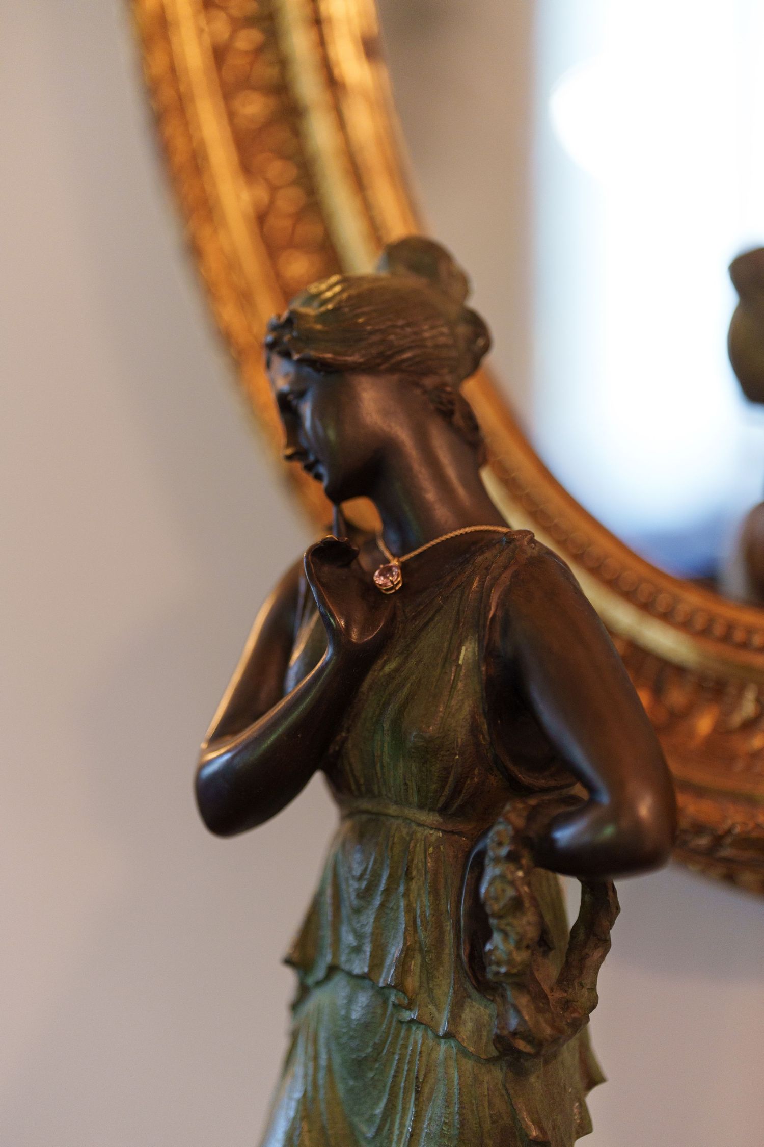 Antique bronze figurine of a classical woman in a flowing dress, displayed in front of an ornate gold mirror.