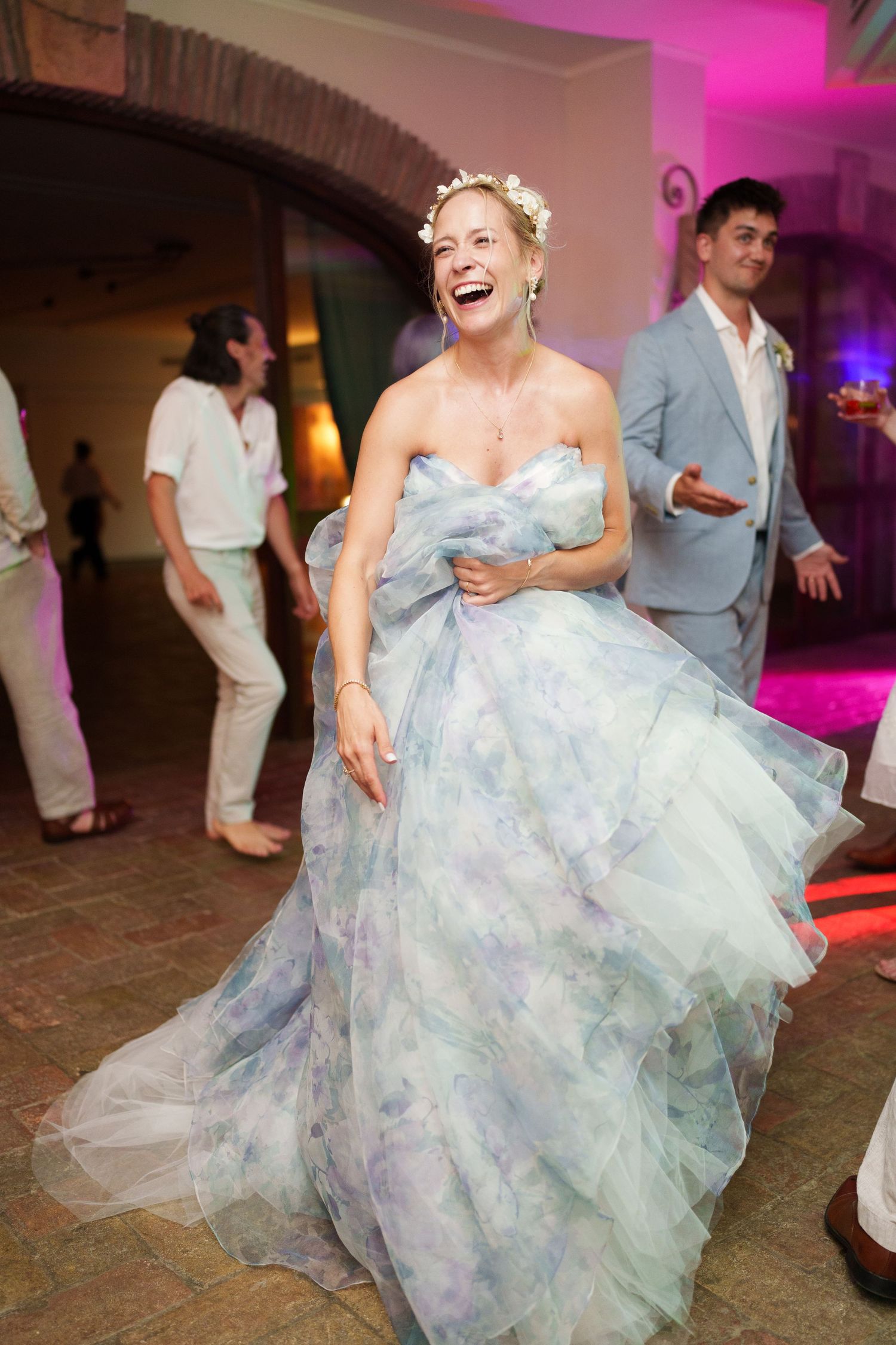 Bride in floral blue tulle gown and flower crown laughing joyfully while dancing at wedding reception.