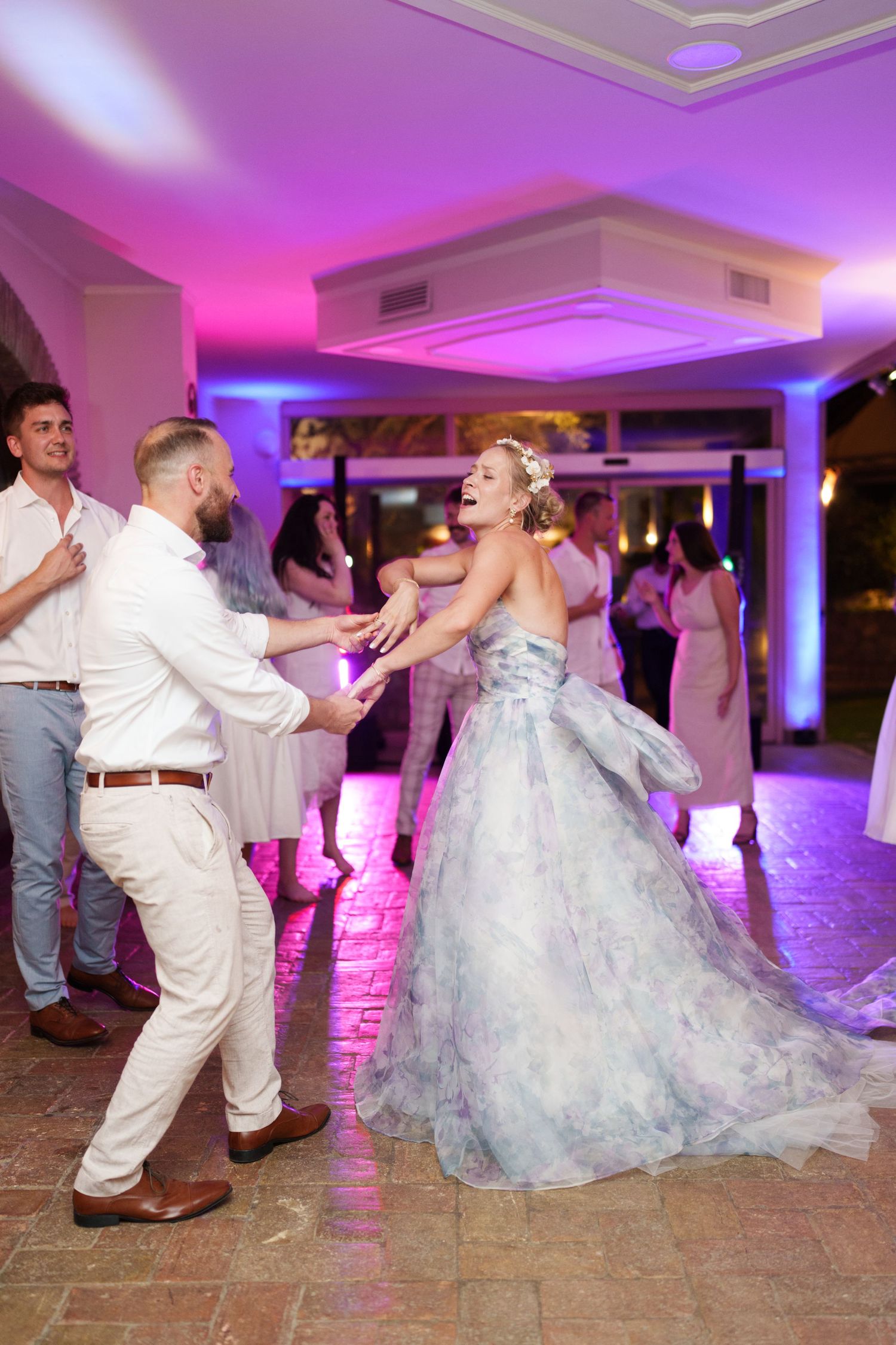 Bride in floral gown and groom dancing joyfully at wedding reception with colorful purple lighting.