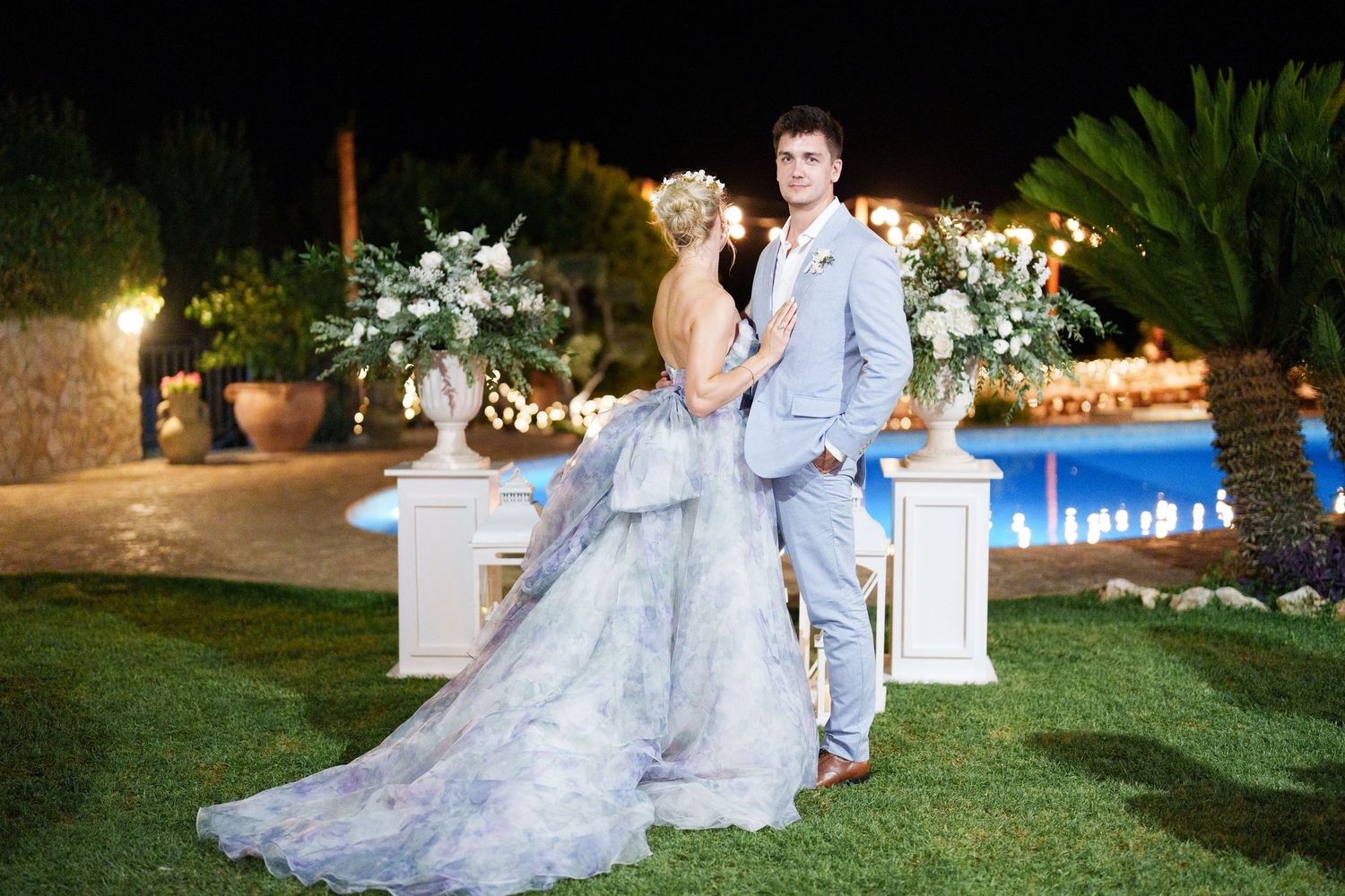 Bride in gray gown and groom in blue suit pose elegantly outdoors at night near a pool with floral arrangements.