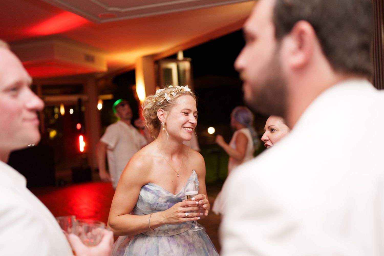 Bride in gray gown smiling at wedding reception with guests in white attire and red ambient lighting.