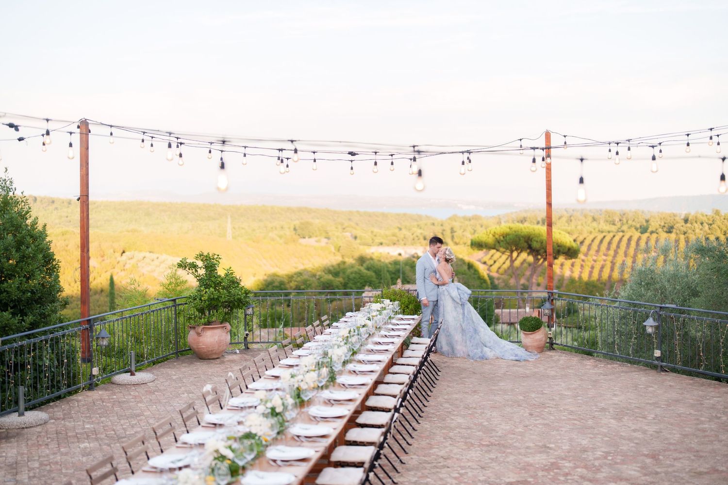 Couple embraces at sunset beside a long elegant outdoor wedding table with string lights and Tuscan vineyard views.