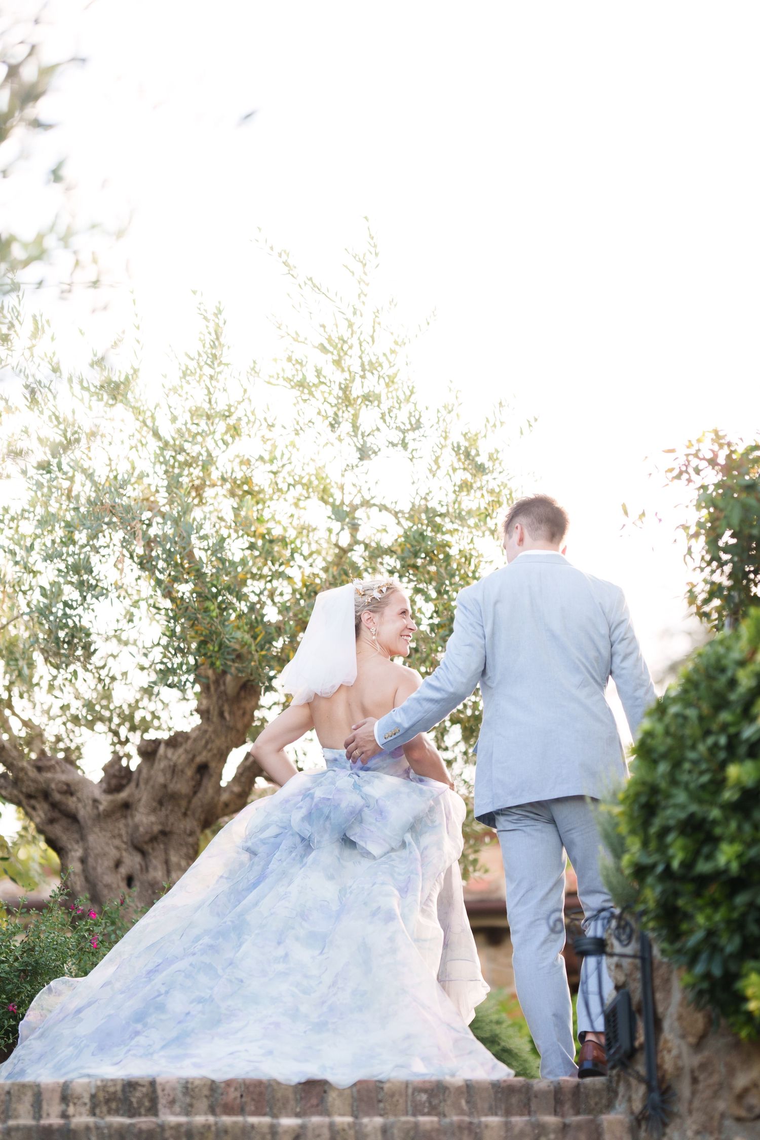 Bride in blue wedding gown and groom in gray suit walk up stone steps surrounded by lush greenery.