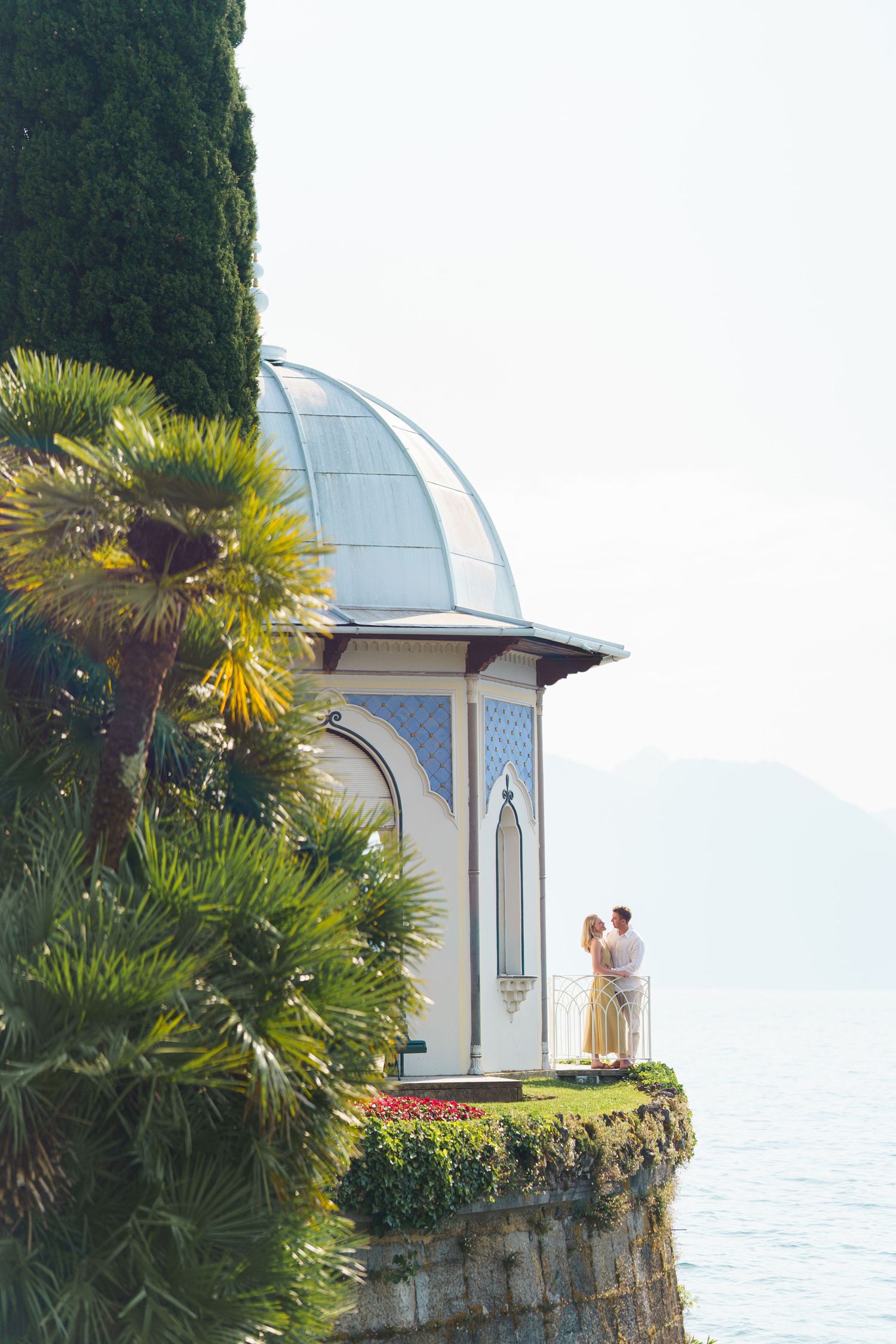 Couple embracing on ornate lakeside pavilion with blue dome, surrounded by palm trees and cypress with mountain views.