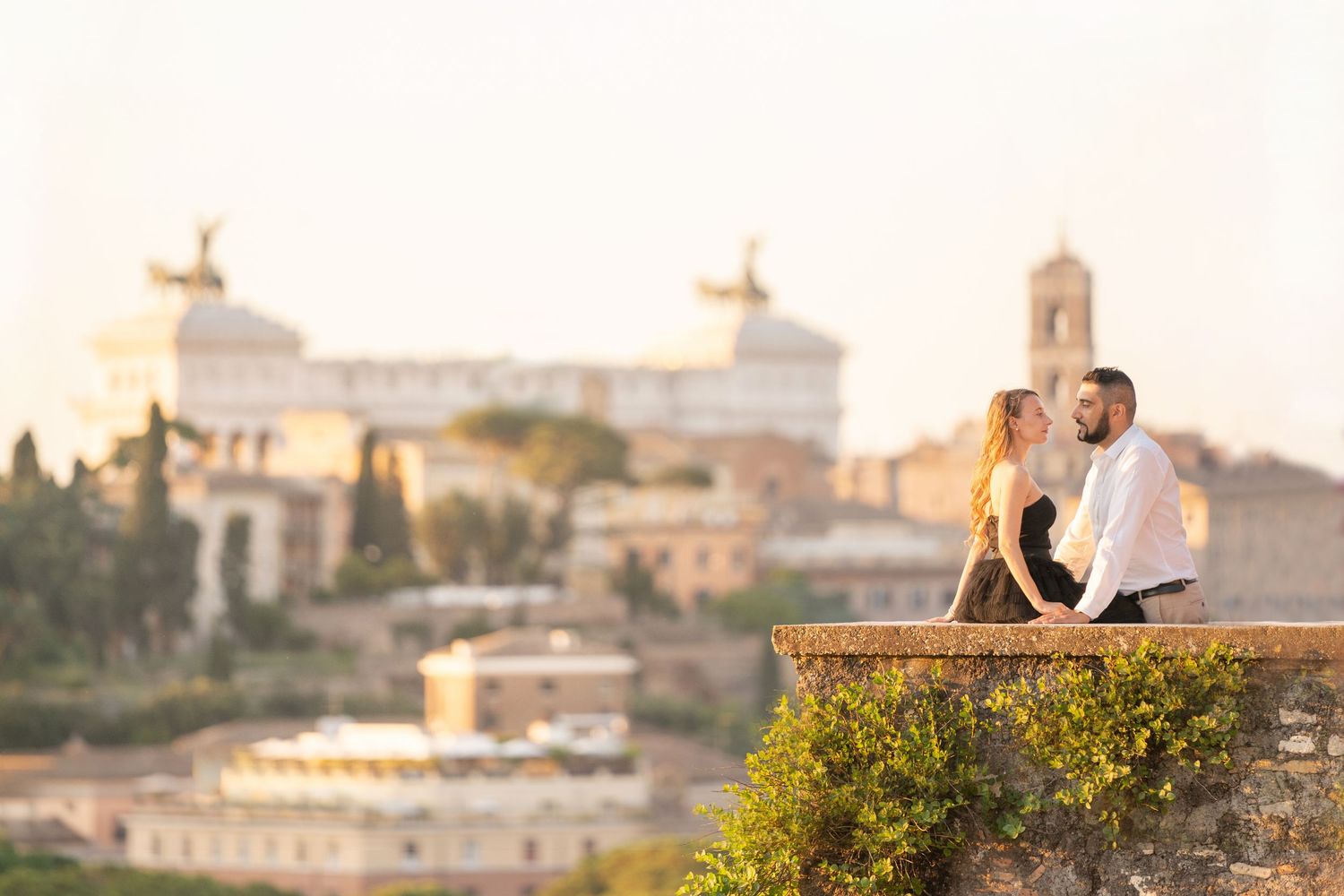 Romantic couple kissing on a stone wall with Rome skyline and Altare della Patria monument at golden hour.