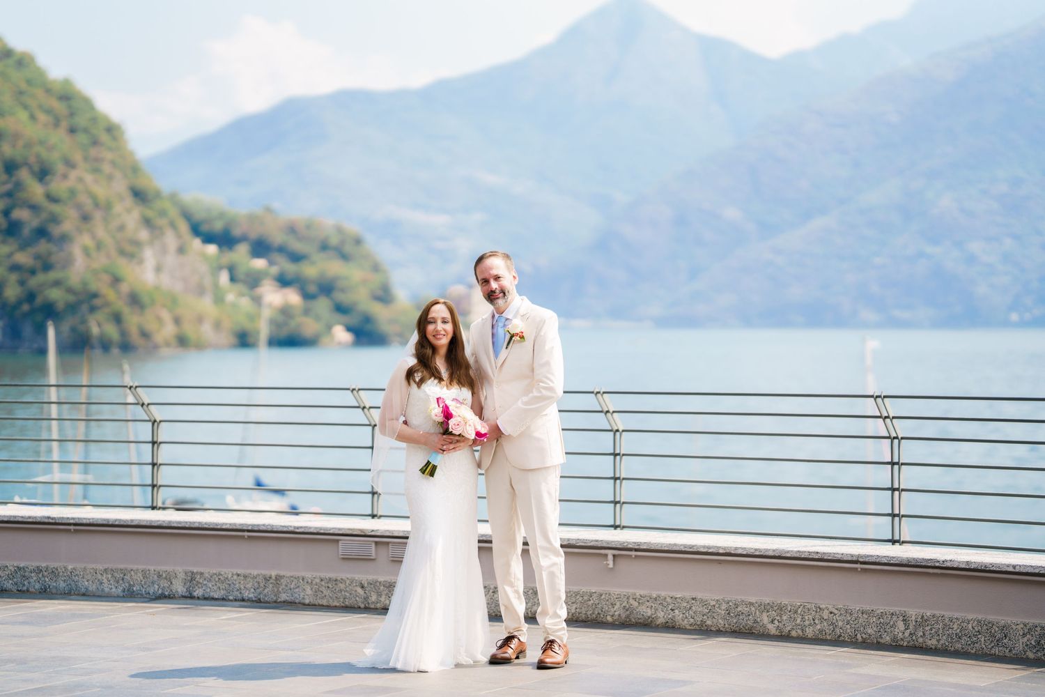 Bride and groom pose on lakeside terrace with mountain backdrop on their wedding day.