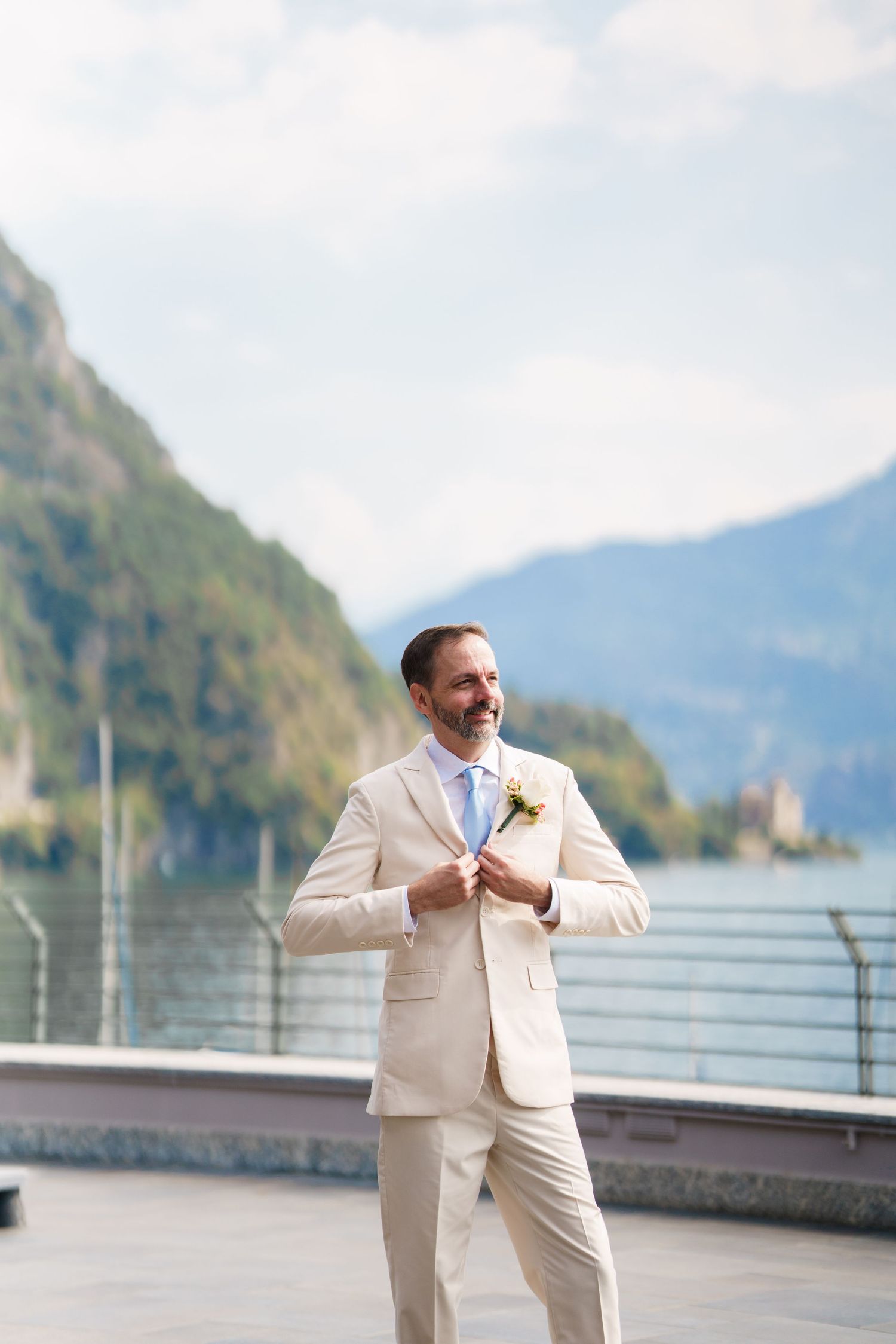 Groom in beige suit with boutonniere adjusting jacket on lakeside terrace with mountain backdrop.