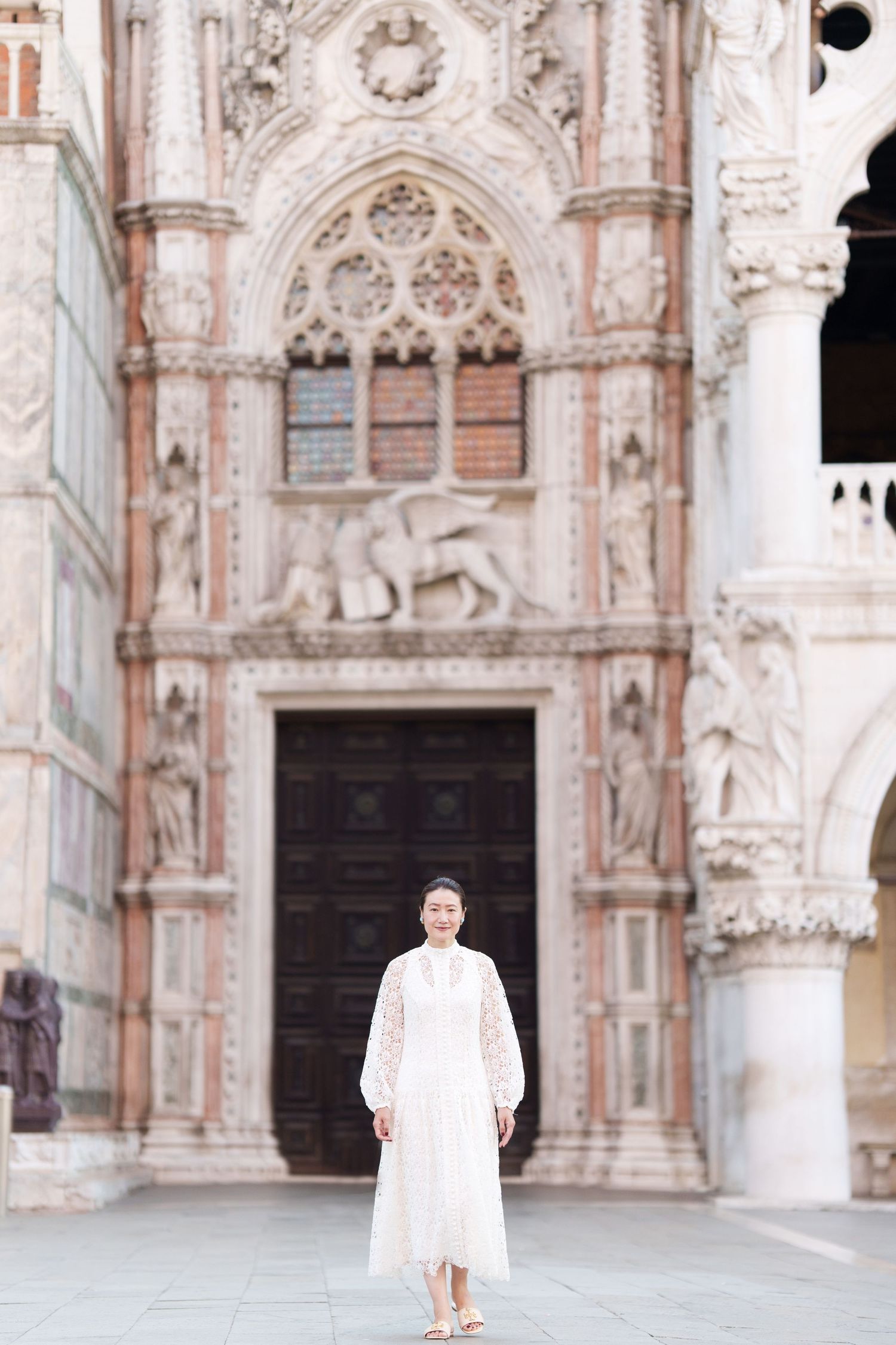 Woman in white lace dress standing before ornate Gothic facade of Doges Palace in Venice, Italy.