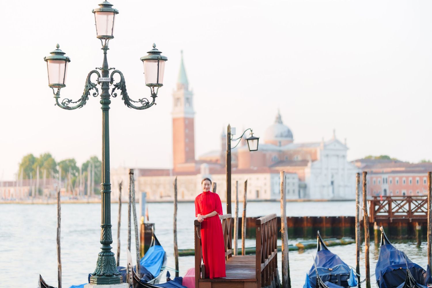 Woman in red dress standing on Venice dock with gondolas and San Giorgio Maggiore church in background.