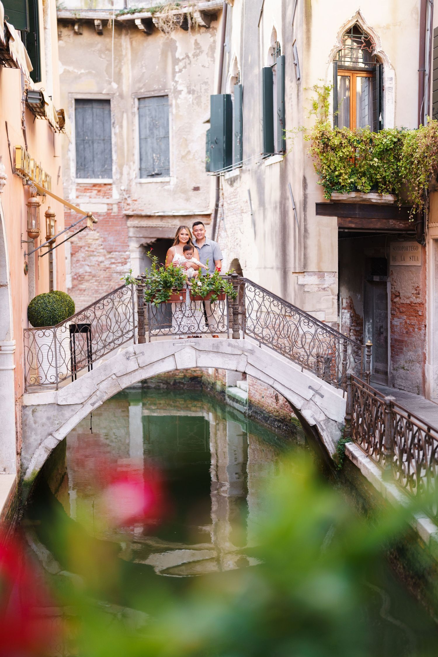 Couple standing on a romantic stone bridge over a Venice canal, surrounded by historic buildings and lush greenery.