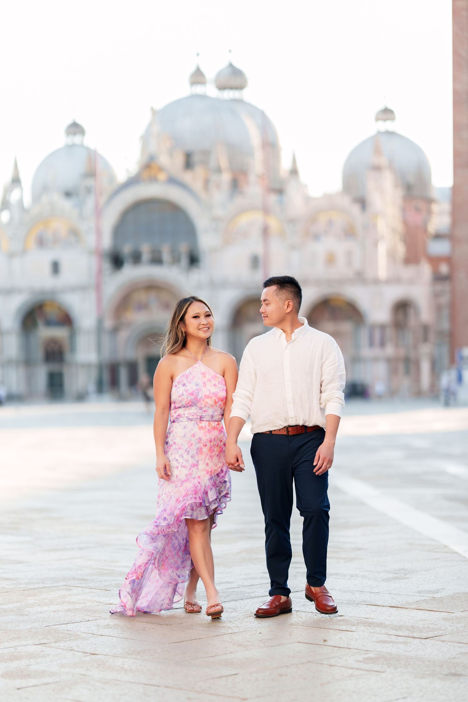 Couple holding hands in front of St. Mark's Basilica in Venice, woman in pink dress, man in white shirt.
