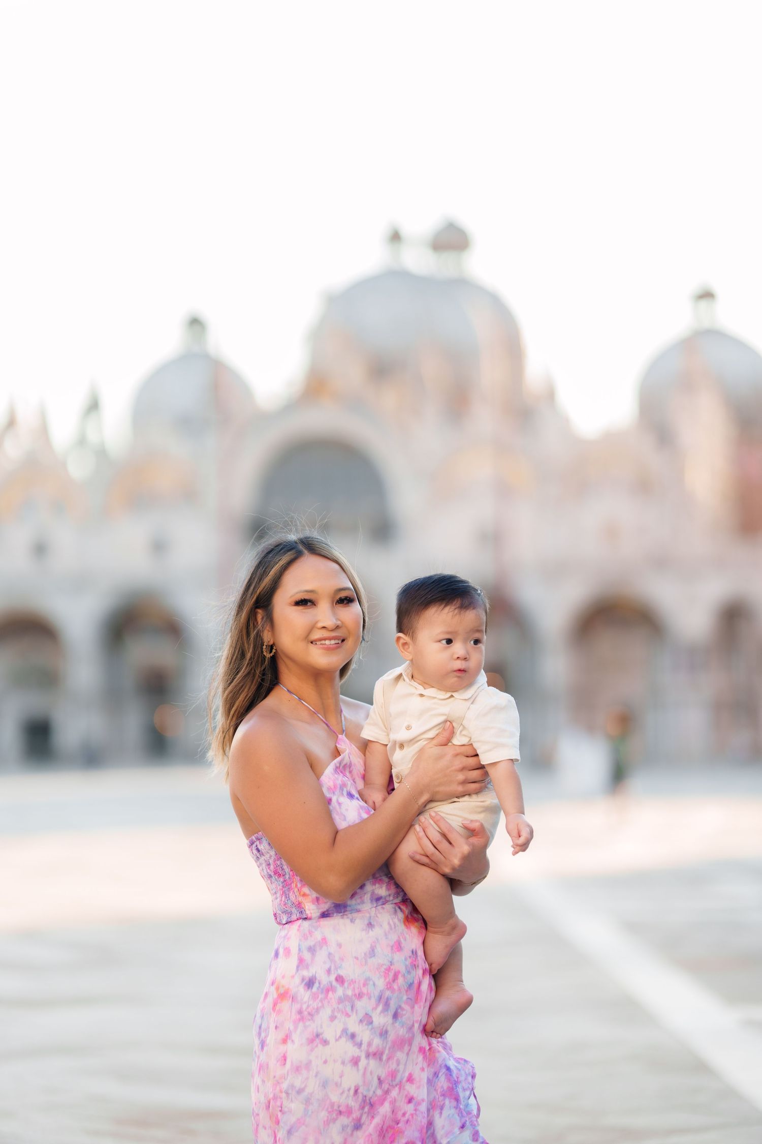 A woman in a floral dress holds her baby in St. Mark's Square, Venice, with the iconic basilica in the background.