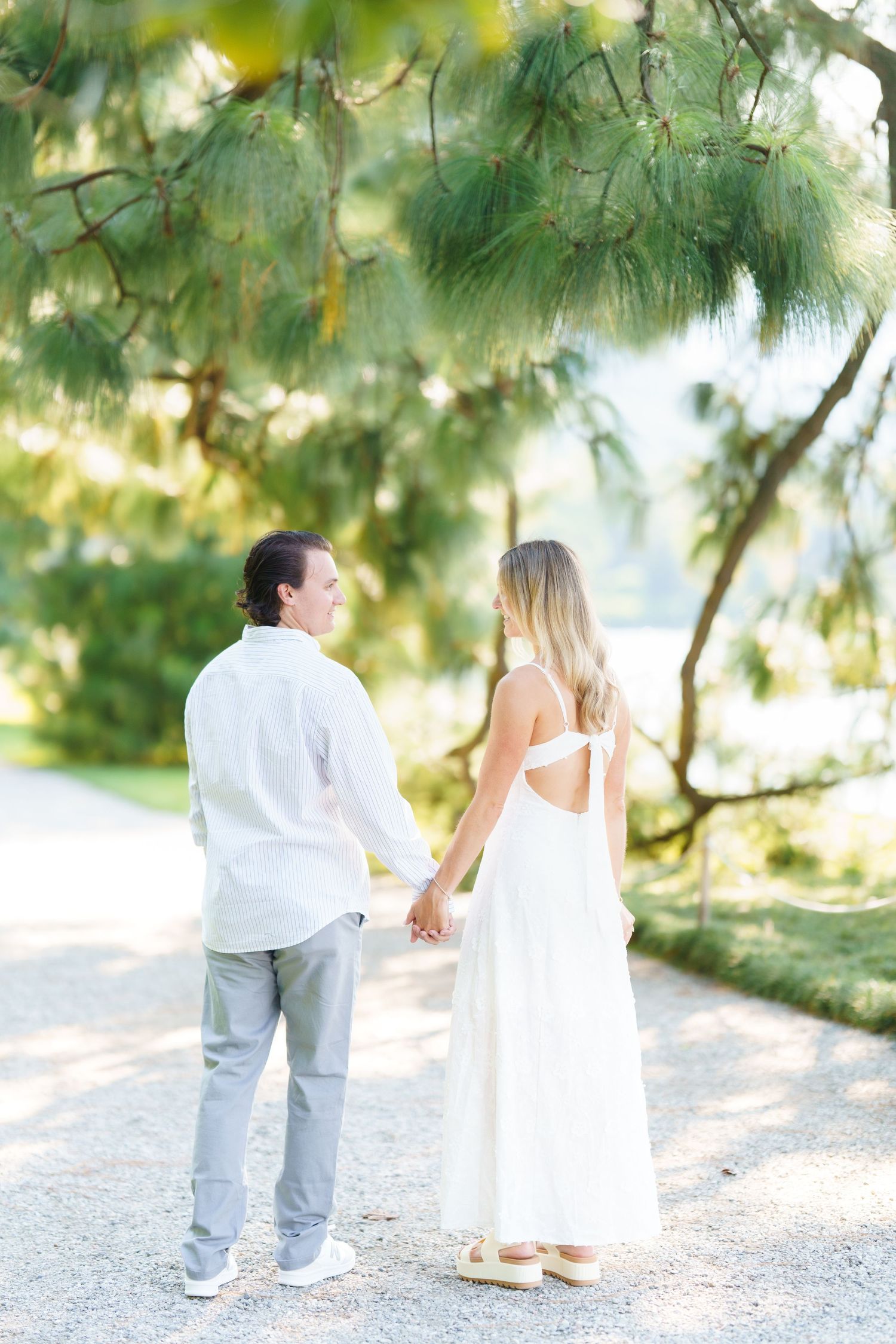 A couple in white and gray attire walks along a gravel path surrounded by green pine trees in a romantic outdoor setting.