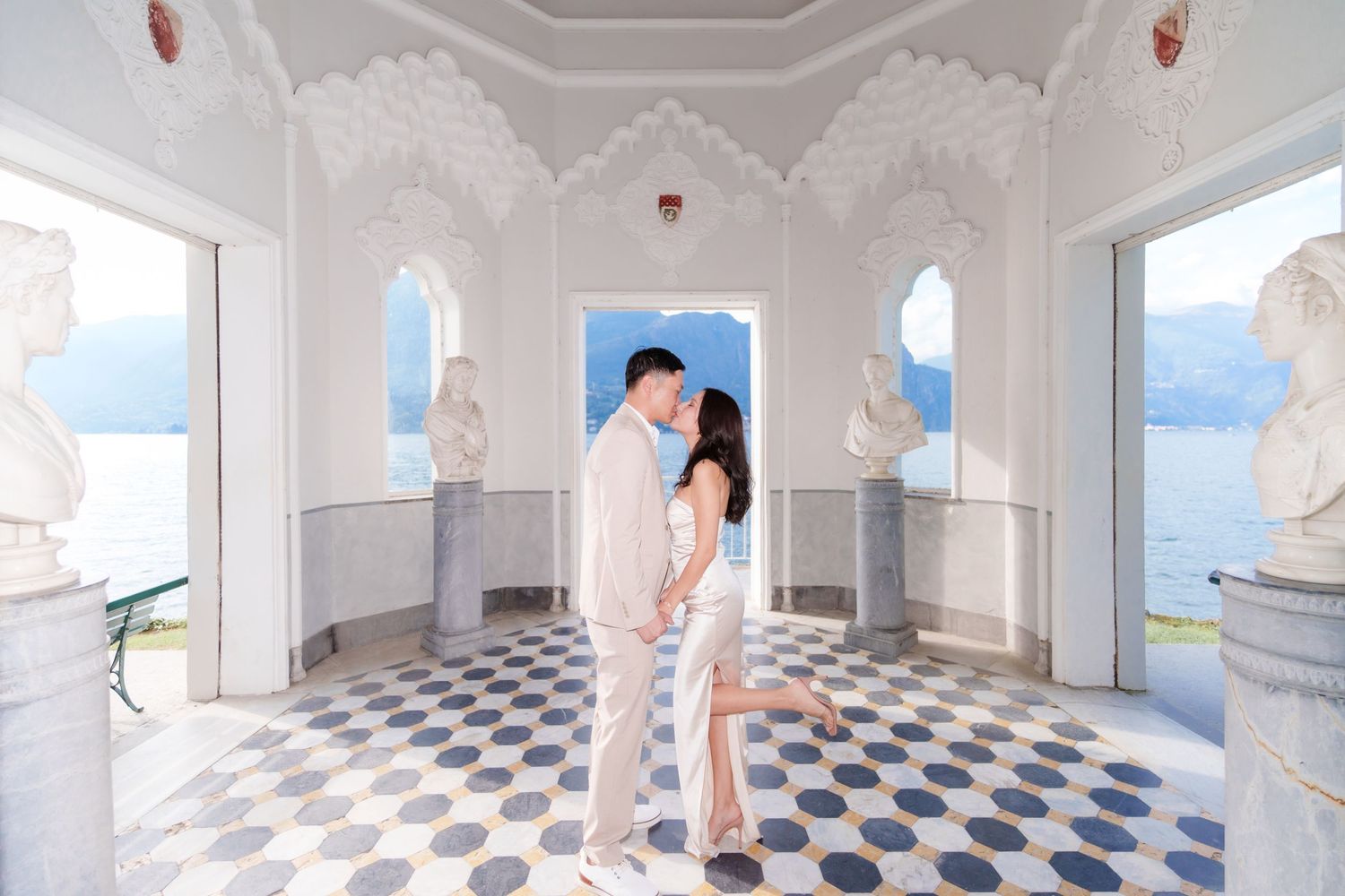 A couple shares a romantic moment in an ornate white palace corridor with checkerboard floors and arched windows.
