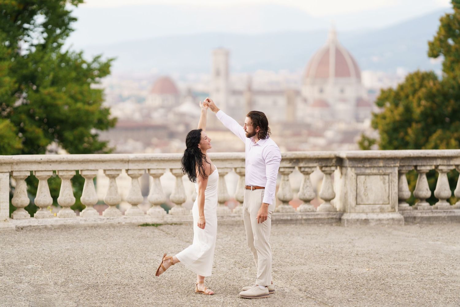 Couple in white outfits walking along a scenic overlook with Florence's Duomo cathedral visible in the background.