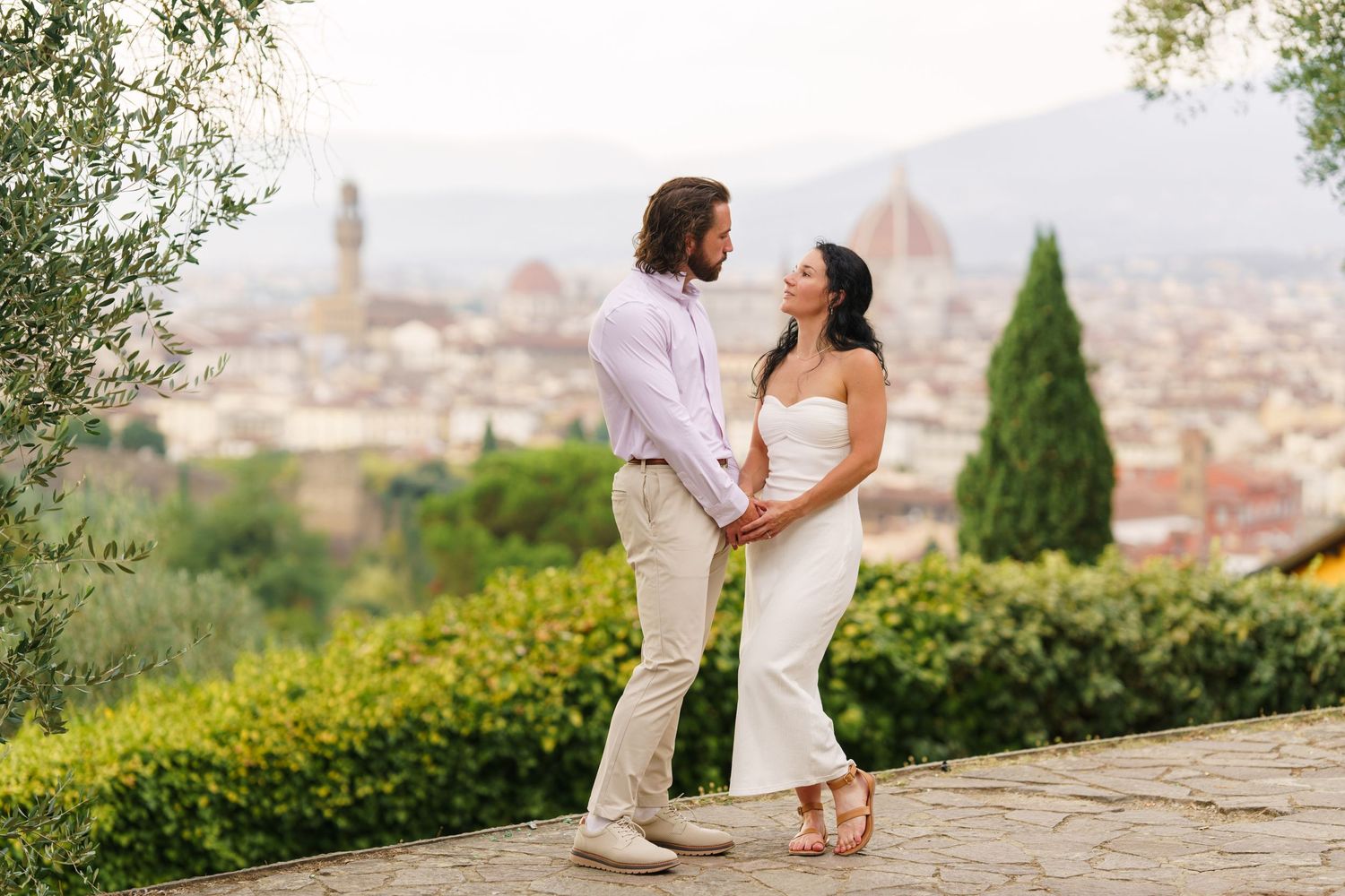 A romantic couple in white outfits embraces while overlooking the scenic cityscape of Florence, Italy from a hilltop garden.