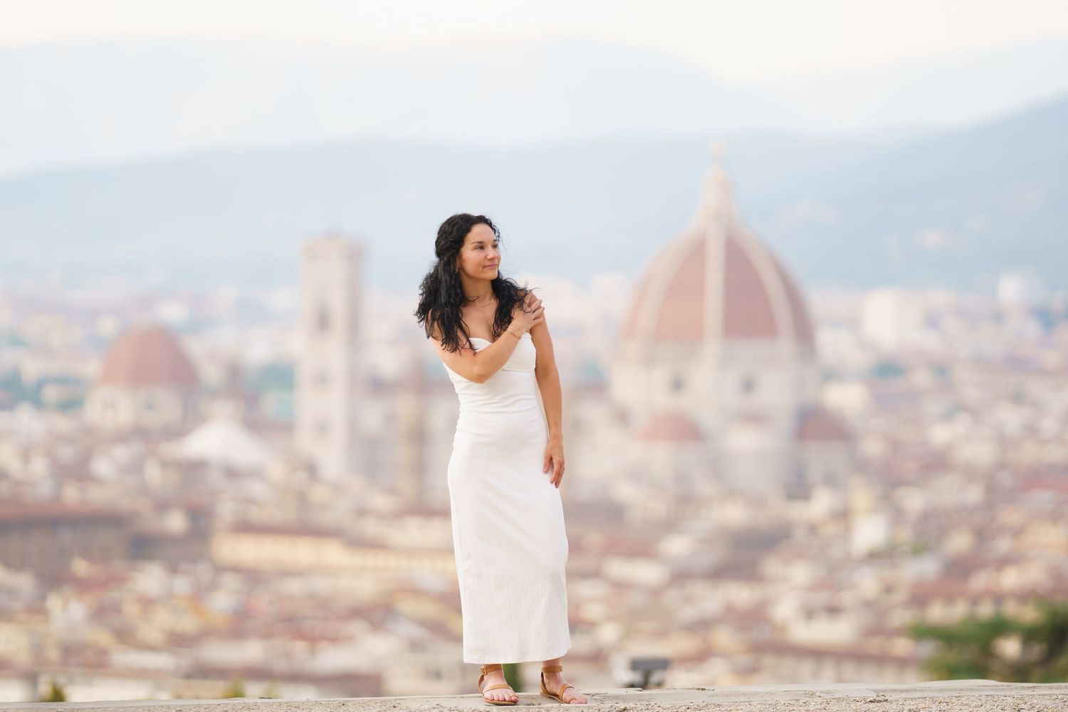 A woman in a white dress stands overlooking Florence, Italy with the iconic Duomo cathedral dome visible in the background.