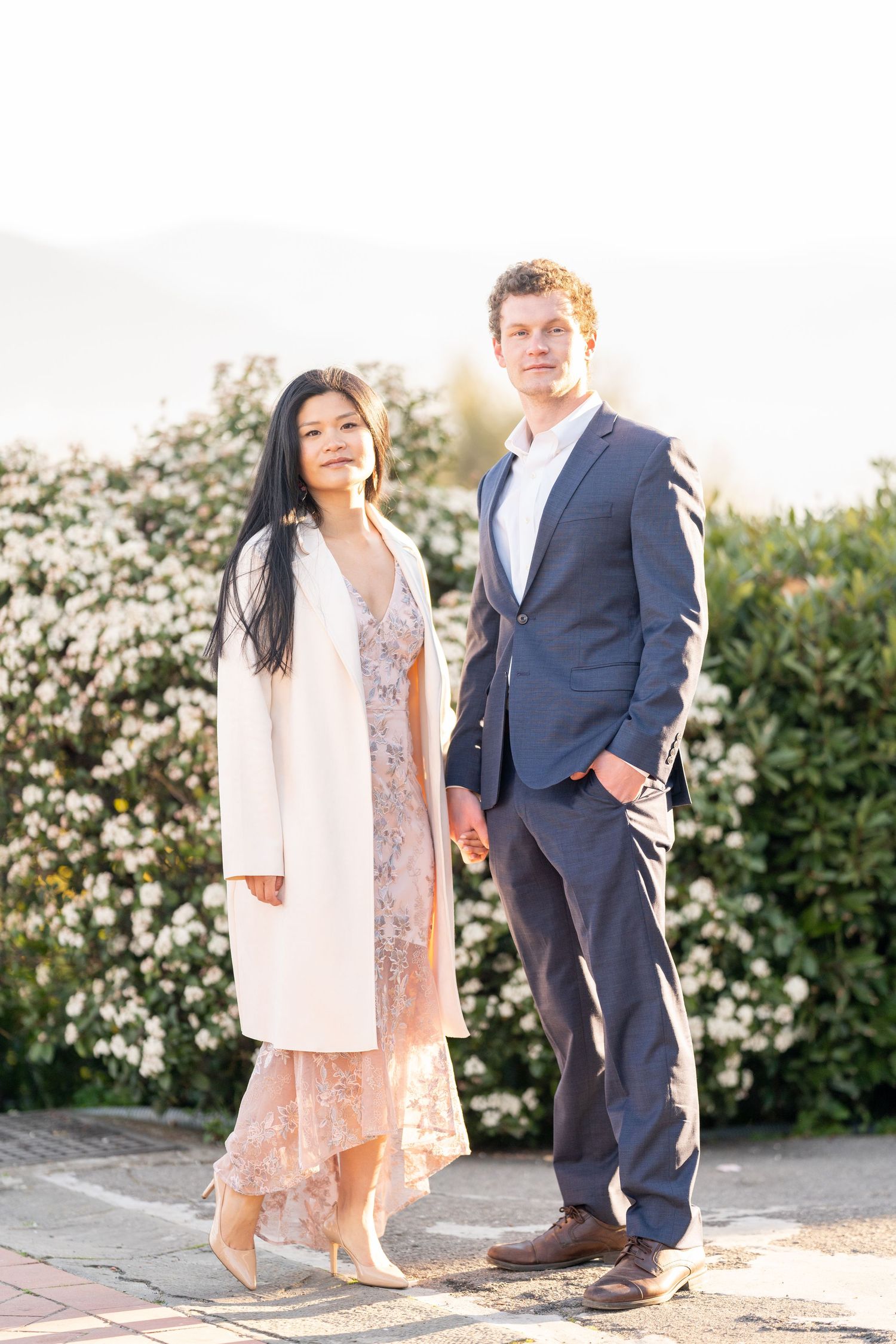 A couple poses together outdoors in formal attire with white flowering bushes in the background during golden hour.