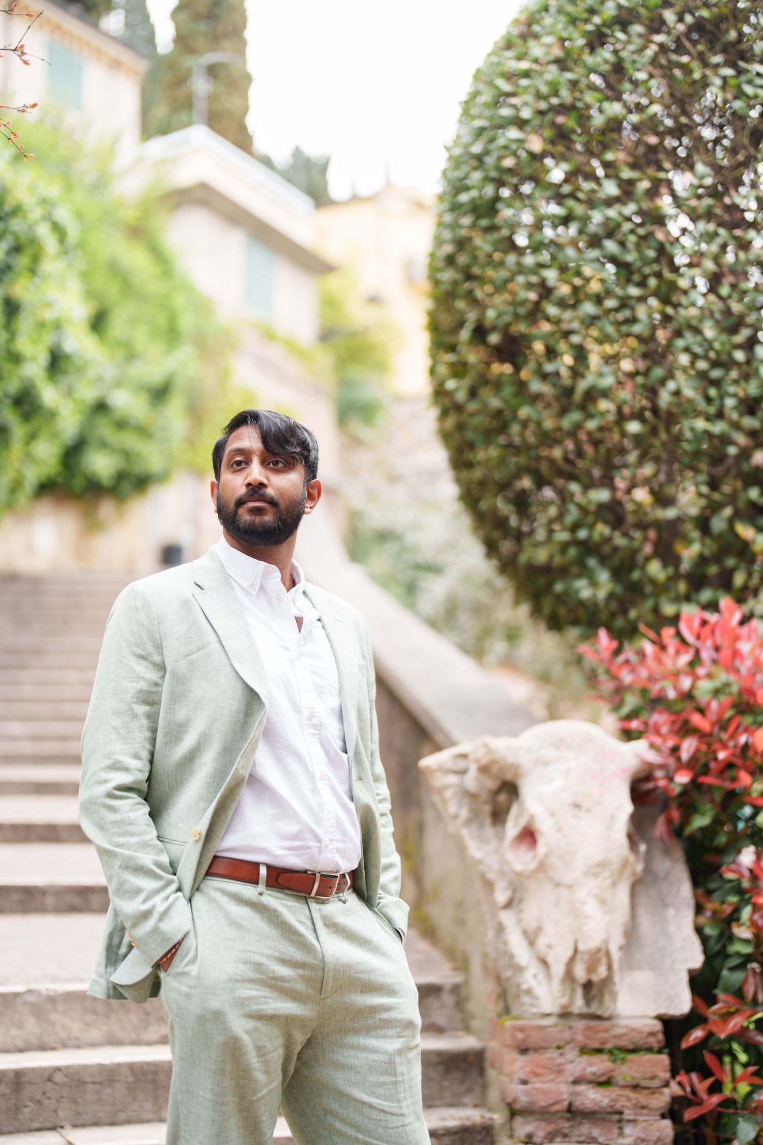 A person wearing a light mint green suit poses on stone steps near flowering plants and shrubs in a garden setting.