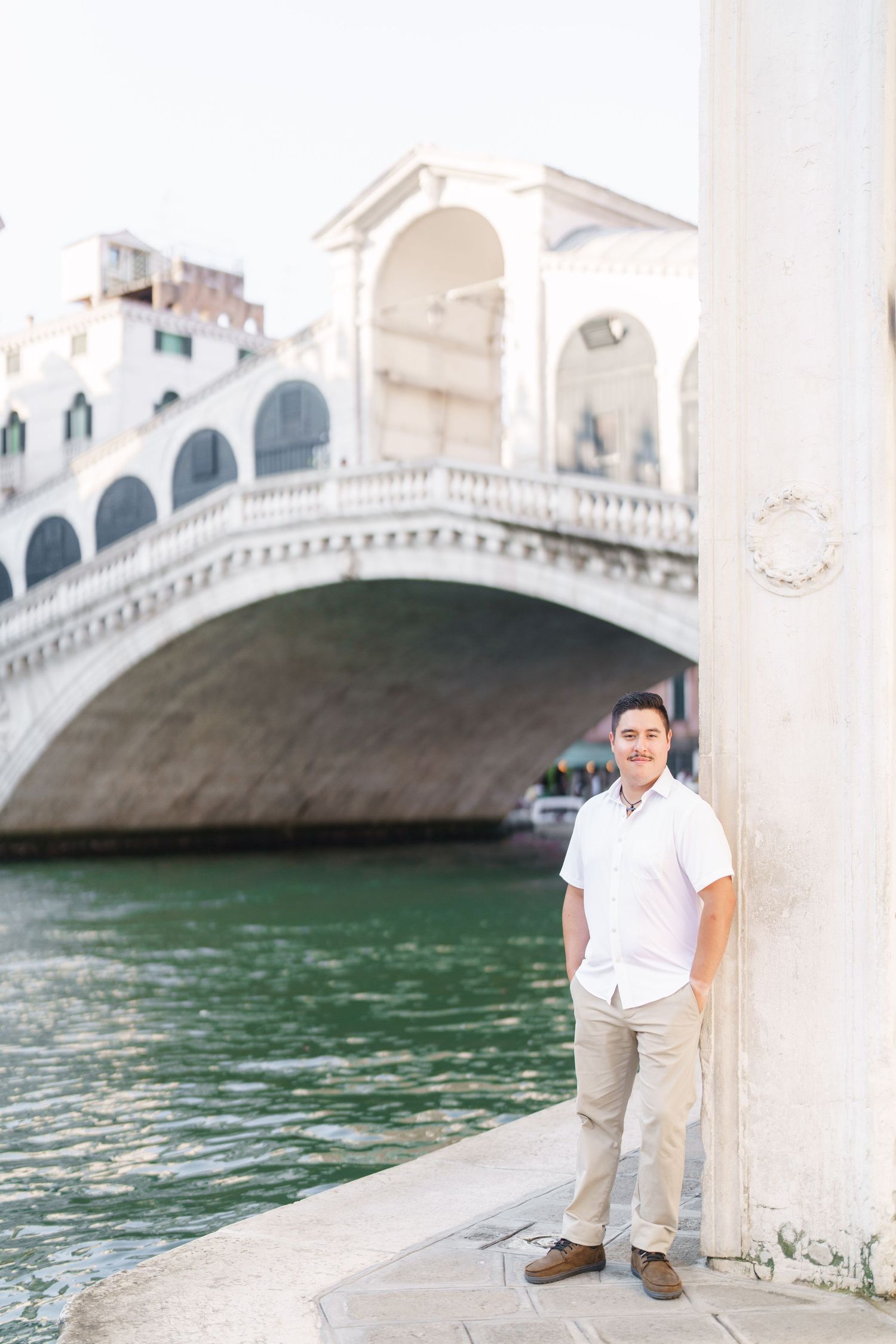 Tourist poses by the iconic Rialto Bridge in Venice, Italy with its white stone arches and canal waters.