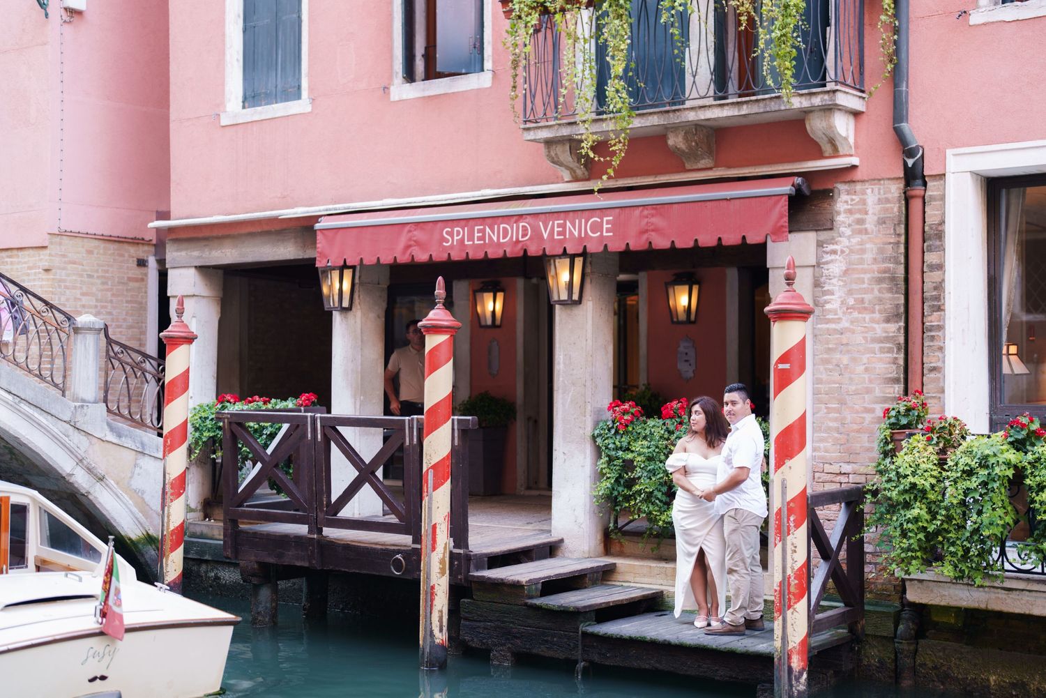 Couple in white robes standing outside a pink spa entrance along a Venice canal with traditional striped mooring poles.