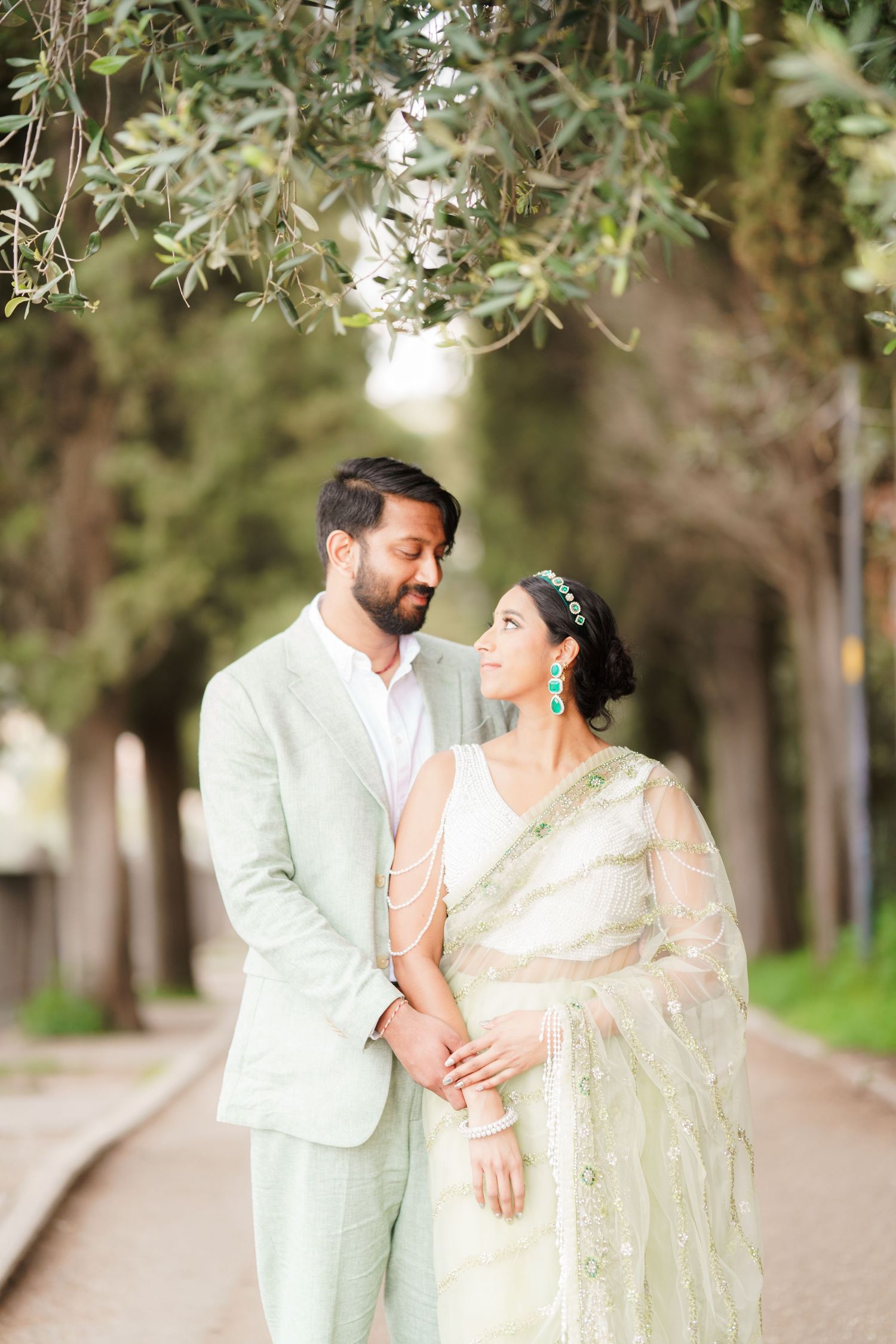 A couple in elegant attire share romantic moments in a tree-lined pathway during a photo session.
