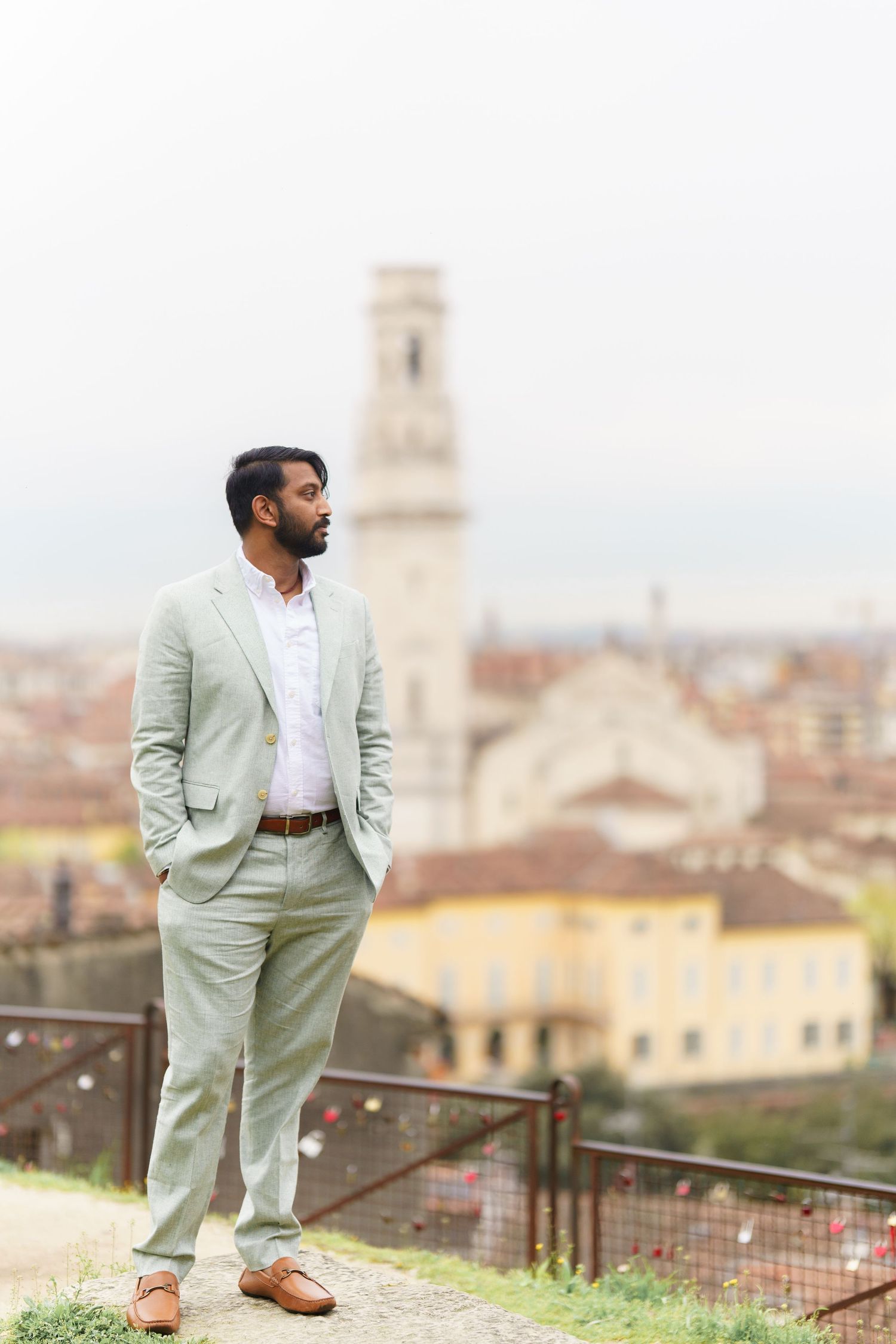 A person in a light sage suit poses on a terrace overlooking a historic European cityscape with church towers.