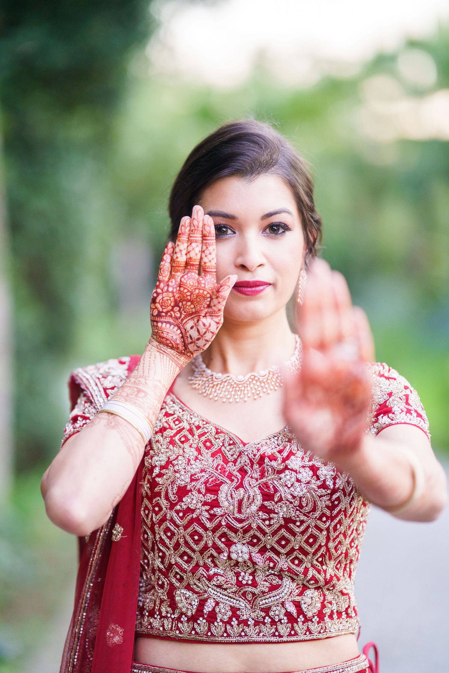 A traditional Indian bride in a red lehenga shows her intricate henna designs and jewelry in an outdoor setting.