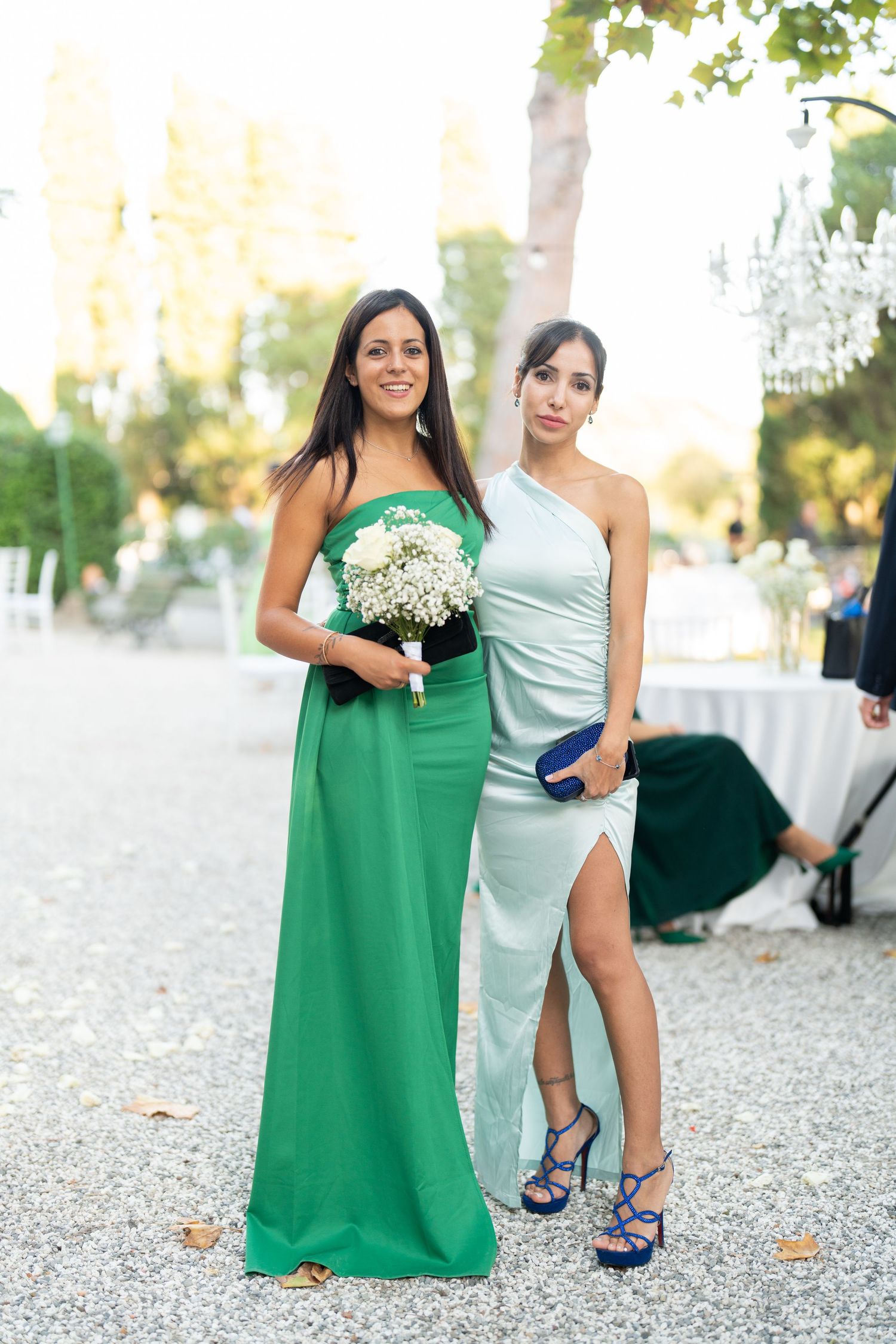 Wedding party members in green and light blue dresses pose together on gravel path.