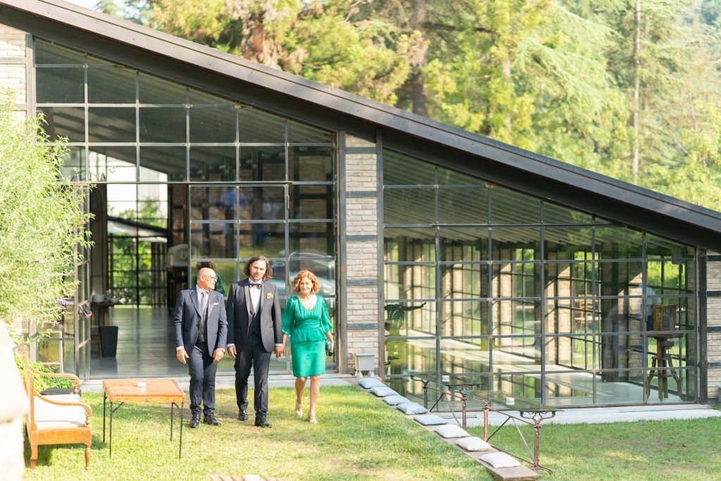 Wedding party gathering outside a modern glass building surrounded by lush greenery on a sunny day.