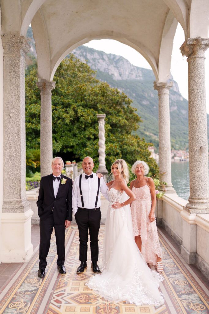 Wedding party poses under ornate archway with lake and mountain backdrop.