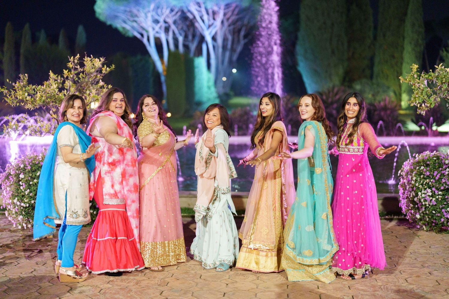 Group wearing colorful traditional Indian dresses poses under purple-lit trees at night.