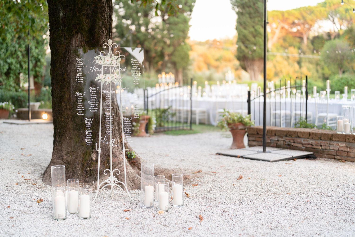 Romantic candle display with crystal chandelier stand under tree at outdoor venue.
