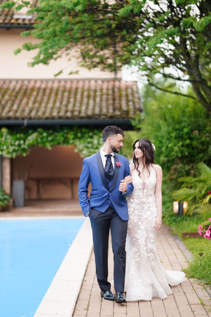 Couple in formal attire walking together on a garden path with lush greenery and traditional architecture in background.