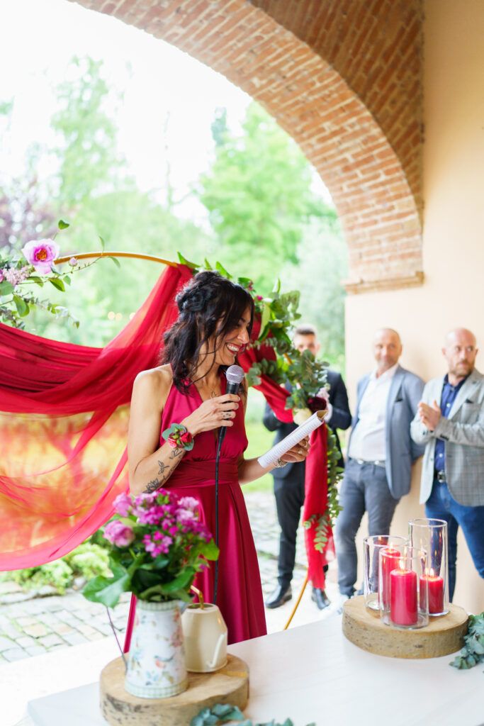 Person in red dress speaking at outdoor wedding venue with brick archway and floral decorations.