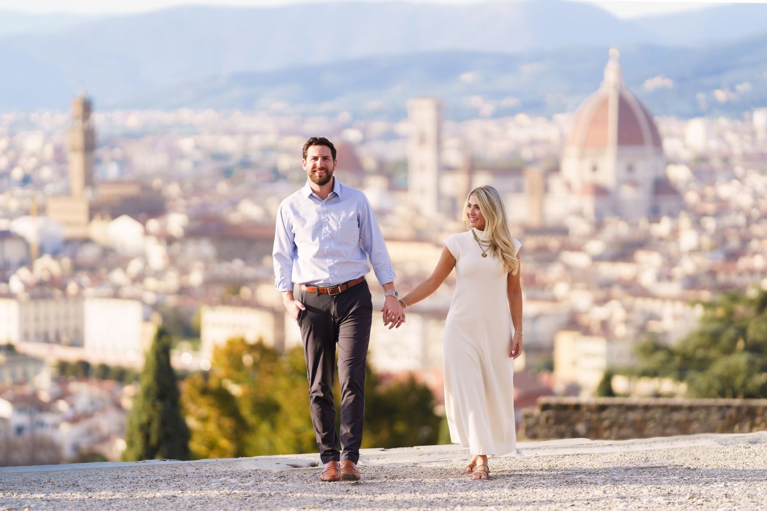 A couple holding hands and running along a scenic overlook with a European city skyline in the background.