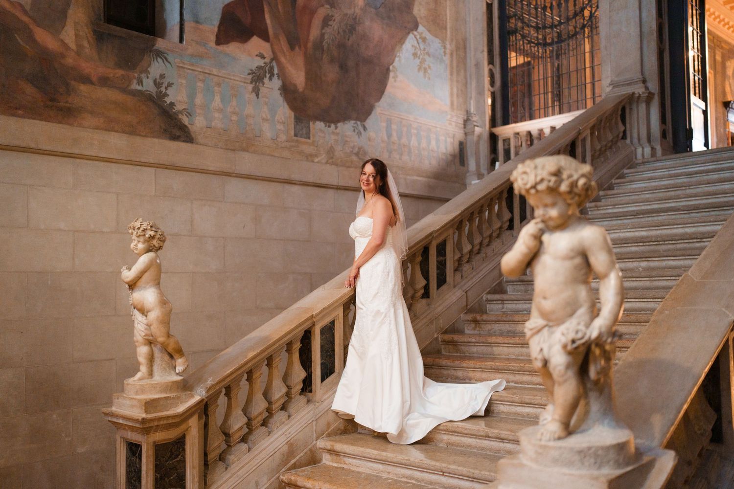 Elegant white wedding gown flowing down ornate marble staircase adorned with cherub statues in historic palace interior.