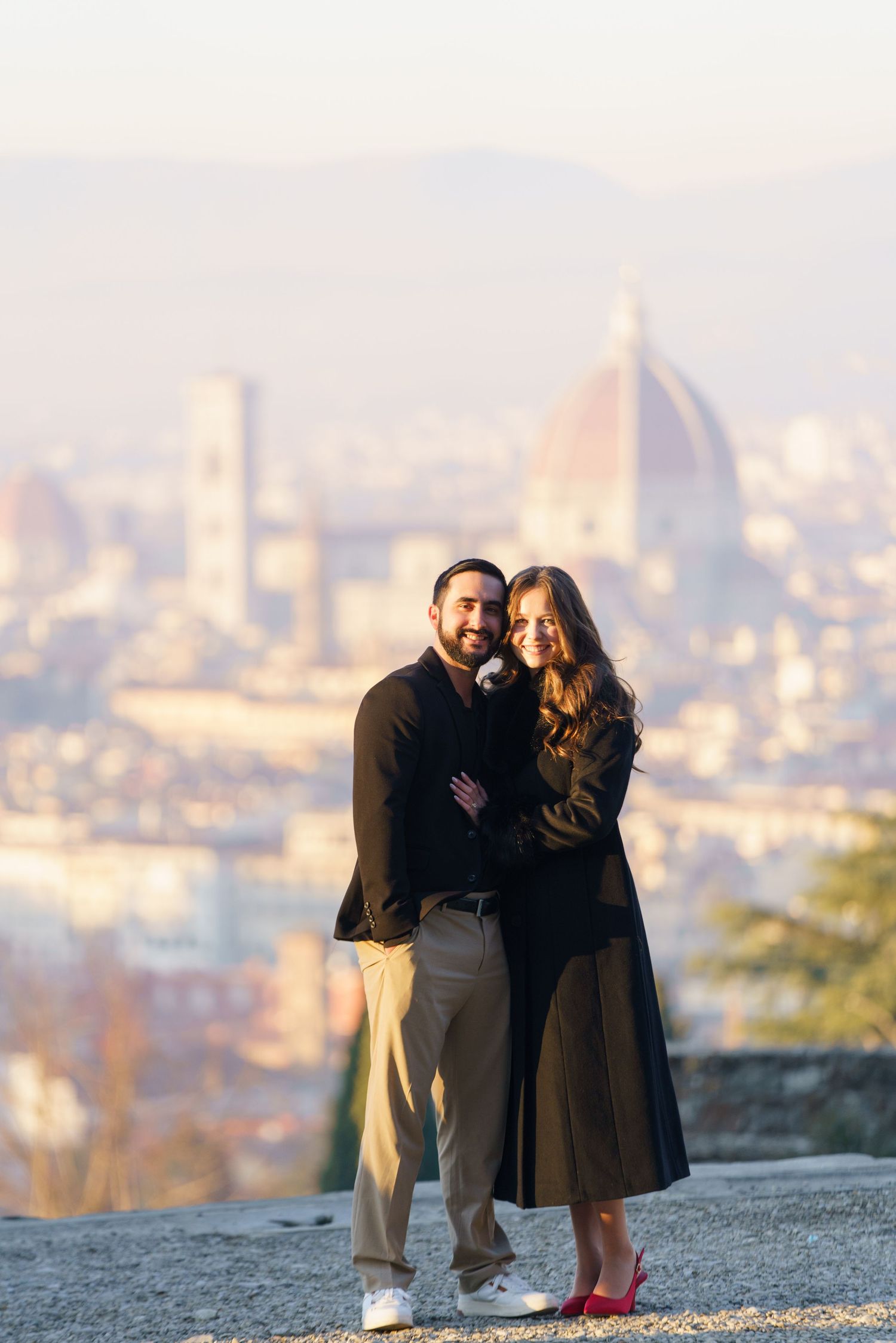 A couple stands together on a hilltop overlooking a city skyline during sunset, creating a romantic silhouette.
