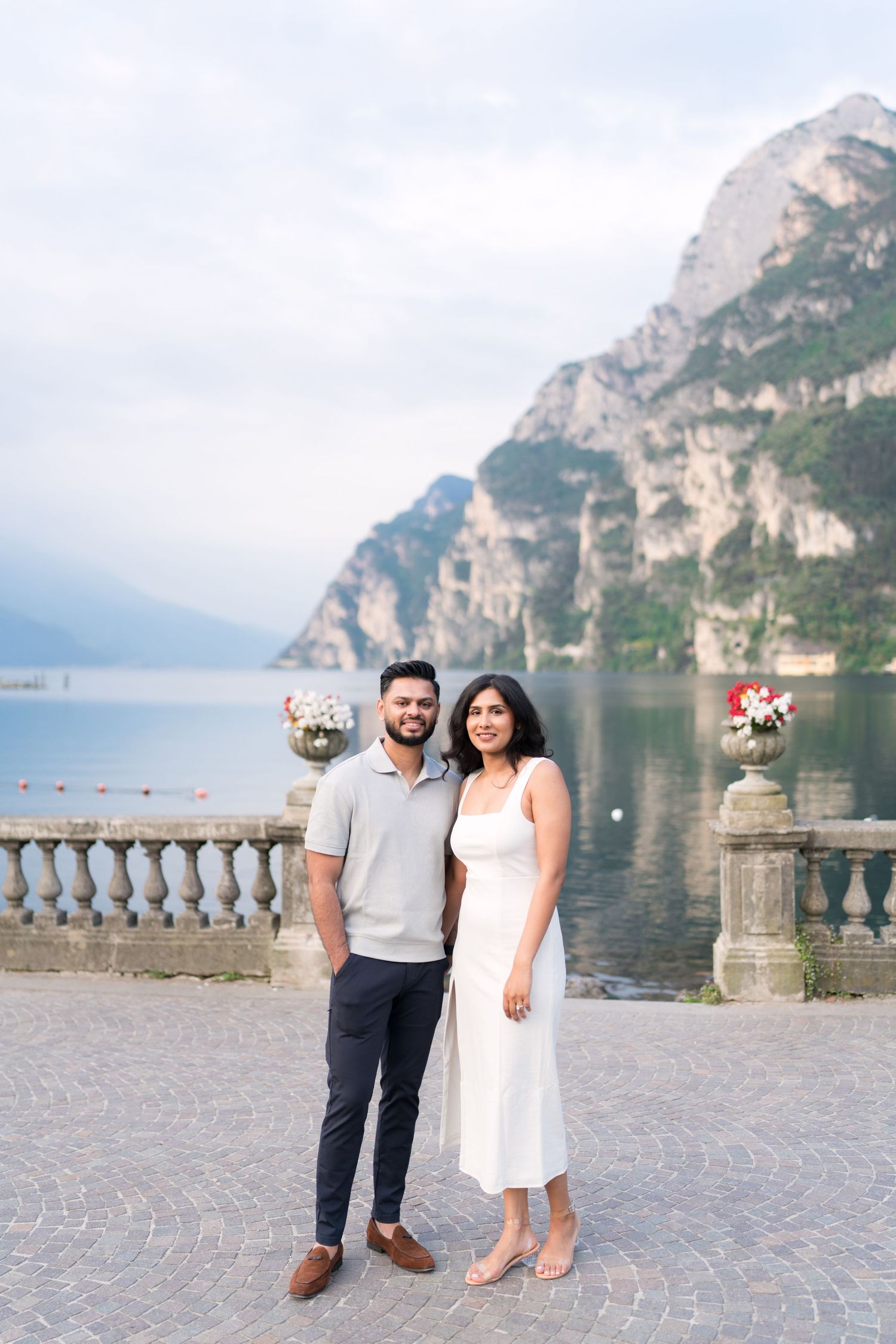 Couple standing on stone terrace overlooking Lake Garda with dramatic mountain cliffs in Italy.