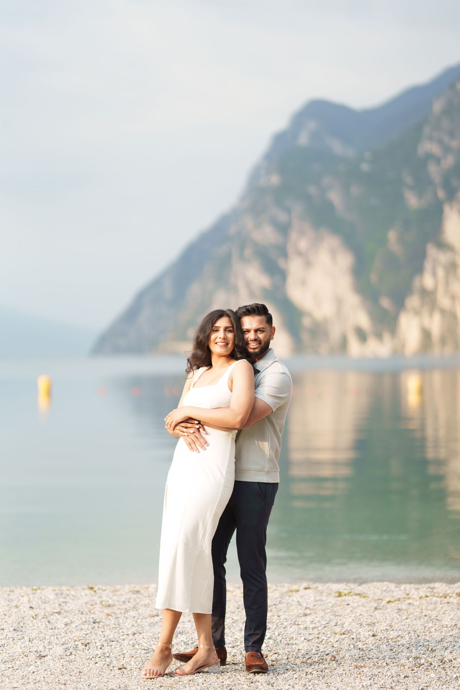 Couple in romantic embrace stands on pebble beach with stunning mountain lake vista in background.