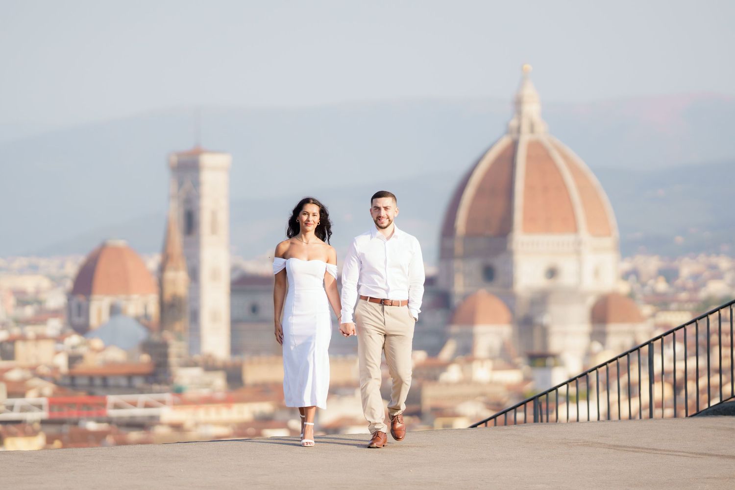 A couple walks along a scenic viewpoint overlooking the iconic Duomo cathedral and Florence skyline at sunset.