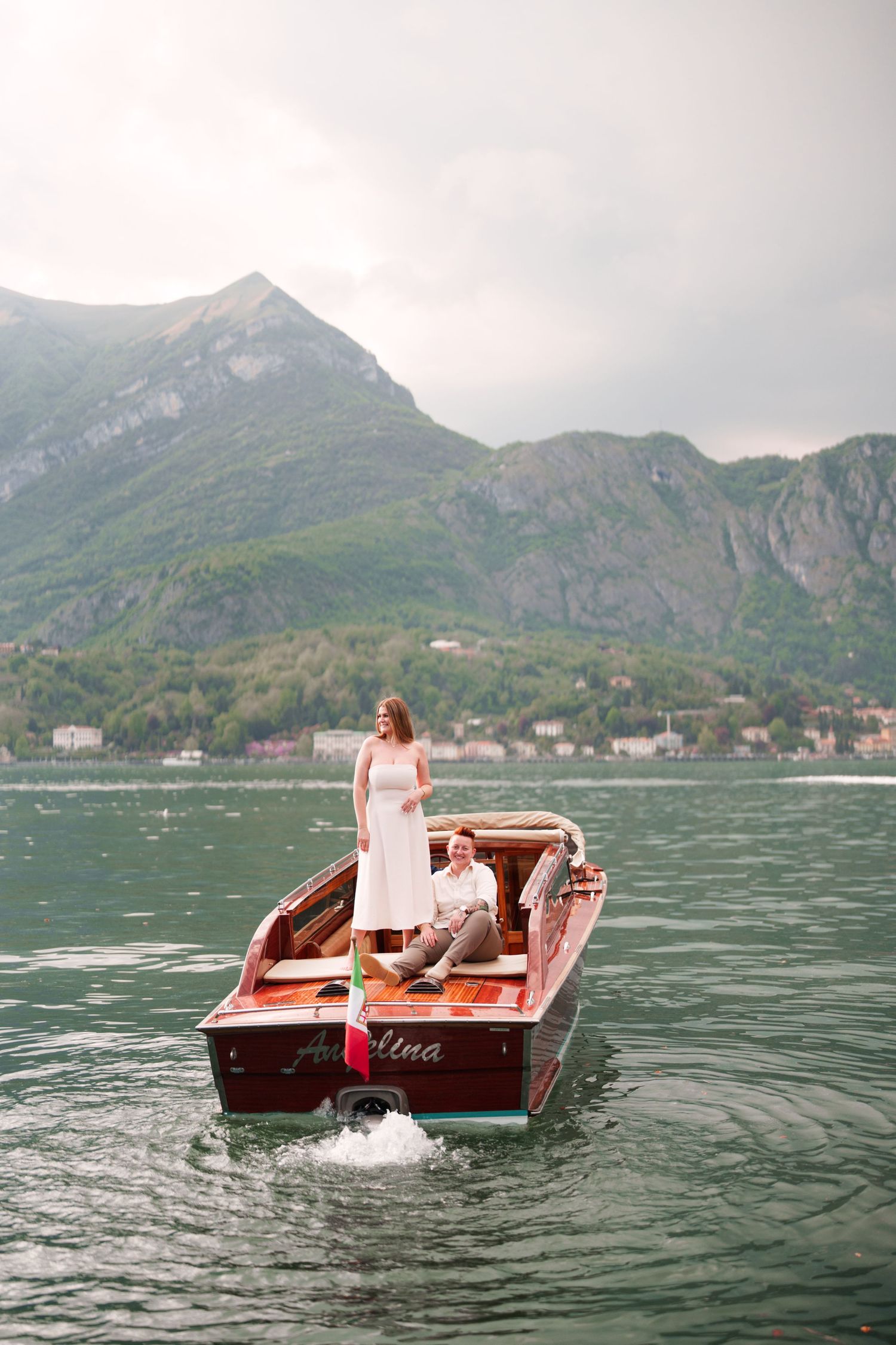 Couple on a vintage wooden boat on a mountain lake, bride in white dress standing, groom seated, scenic alpine backdrop.