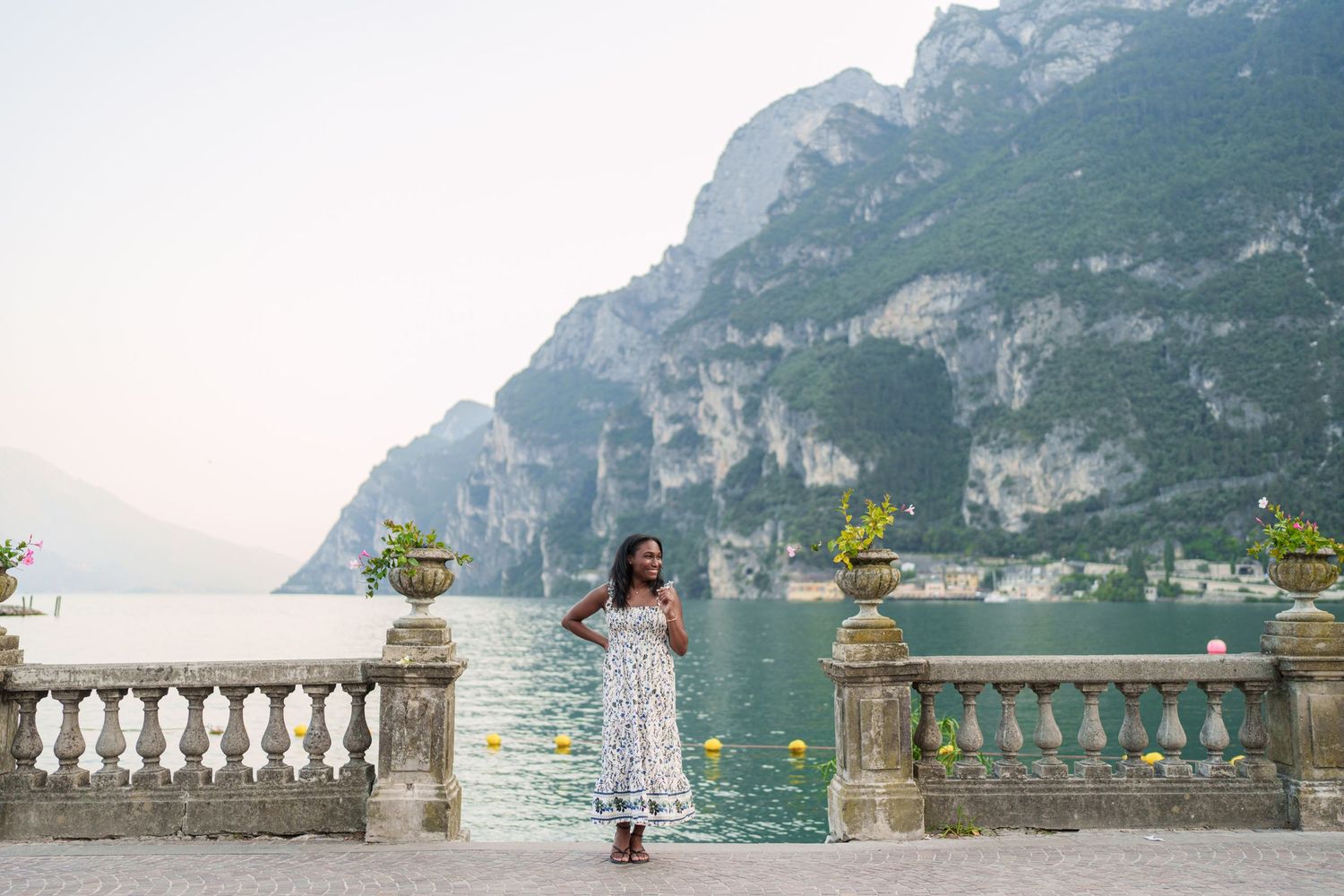 Woman in white floral dress posing on ornate terrace with stunning Lake Garda mountain panorama.