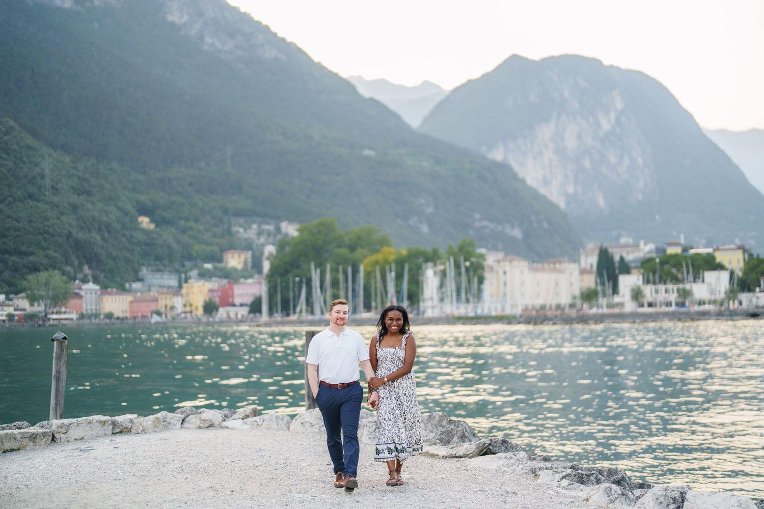 Interracial couple walking hand in hand along a lakeside promenade with mountain scenery.