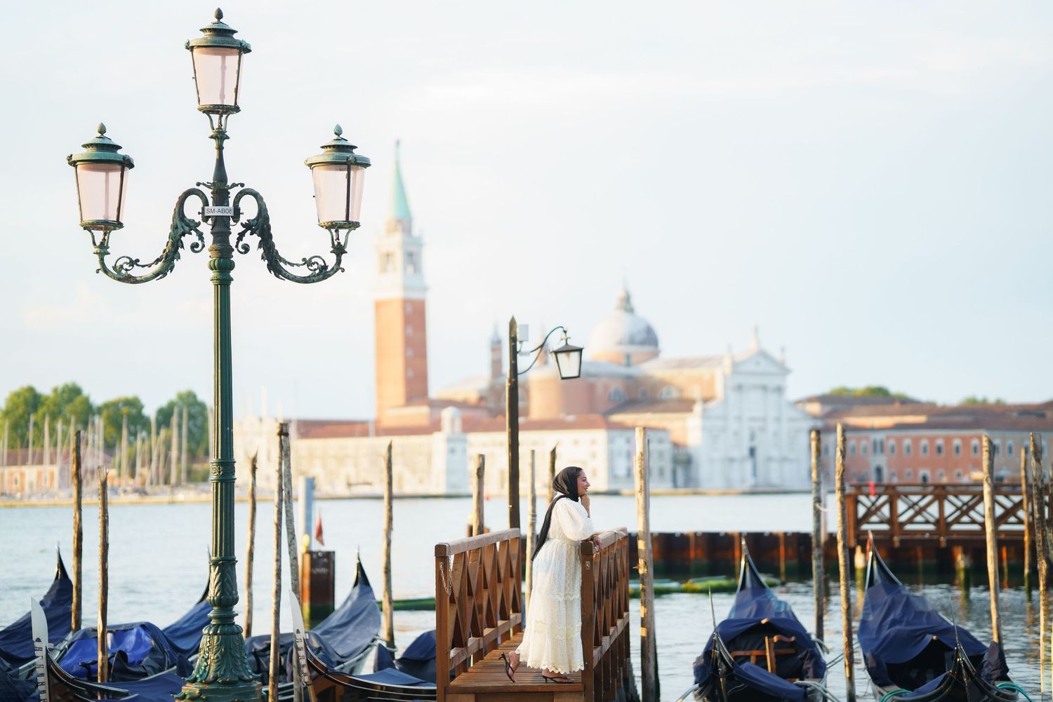 Woman in white dress standing on Venice dock with gondolas and San Giorgio Maggiore church in background.