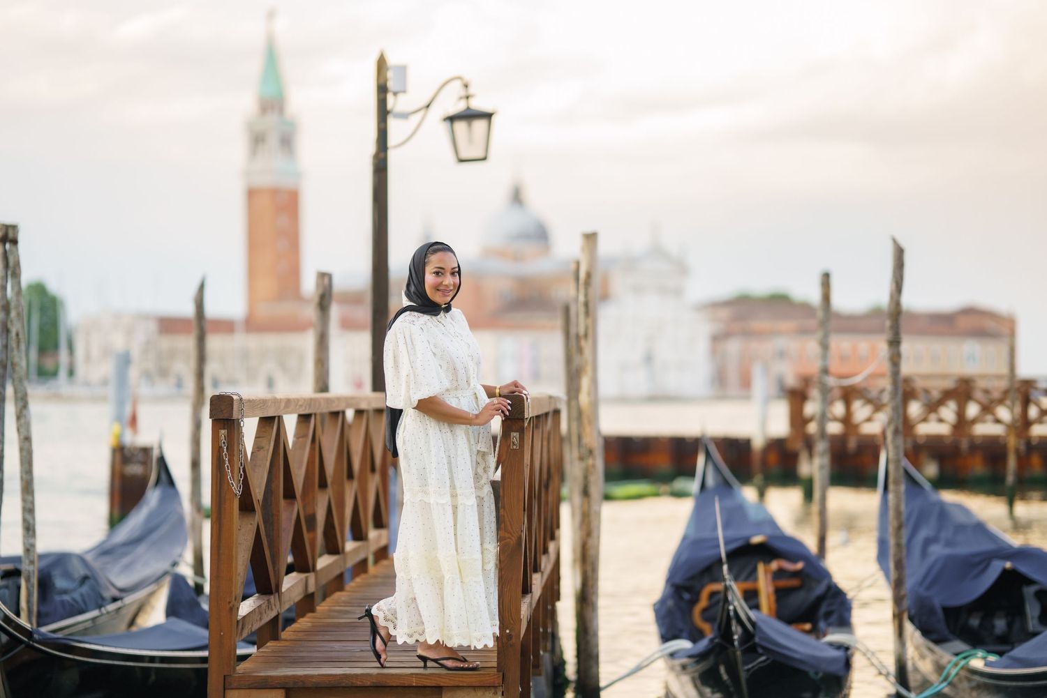 Woman in white dress standing on a wooden dock with gondolas and Venice's San Giorgio Maggiore in the background.