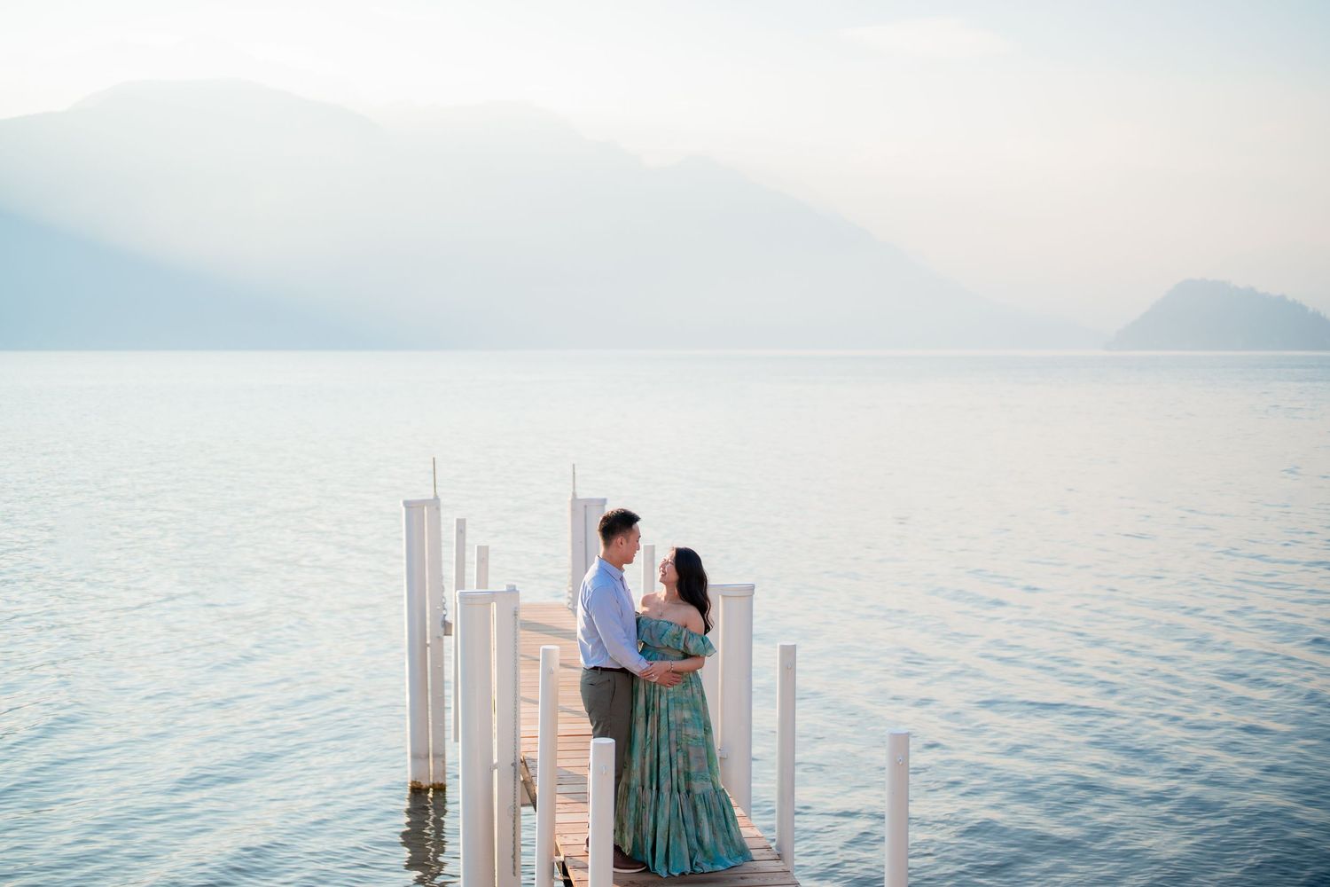 Couple standing on a white dock overlooking a serene lake with misty mountains in the background.