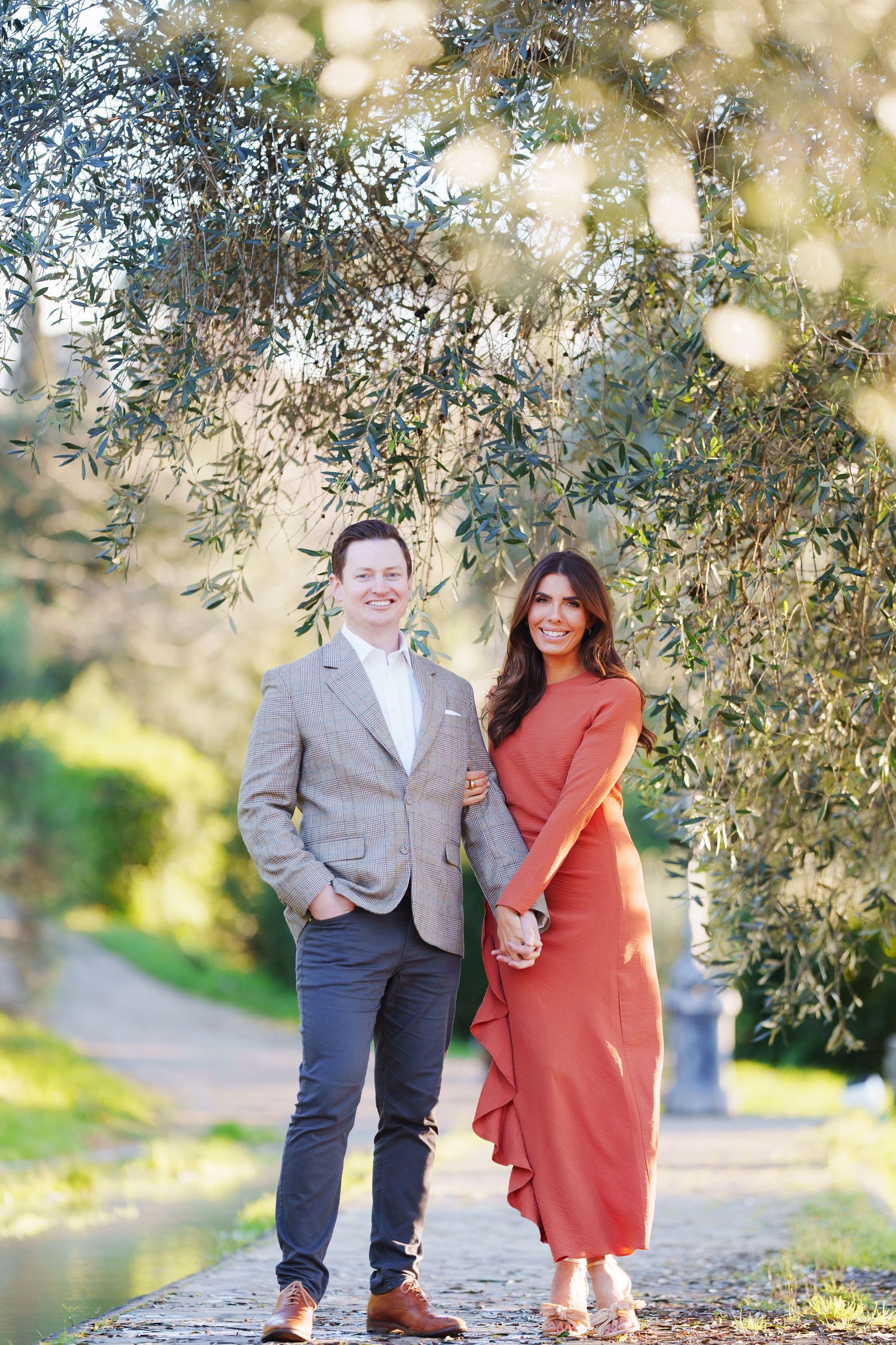 Couple posing outdoors under olive tree branches, man in tweed blazer and woman in rust red dress, garden path background.
